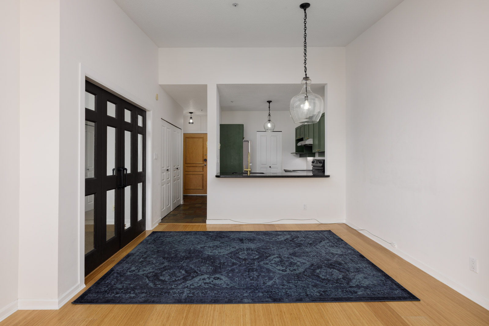 Bright apartment interior with light wood floors, a large blue rug, black-framed double doors, open kitchen with green cabinets, and pendant lights over the counter.
