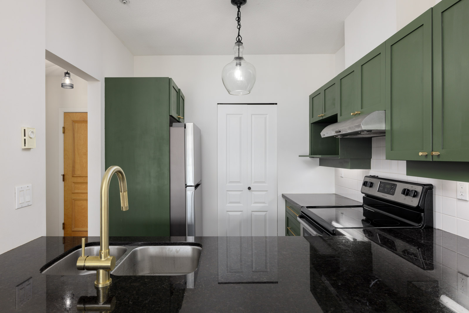 Modern kitchen with green cabinets, stainless steel appliances, a black granite countertop, a double sink with a gold faucet, and a central pendant light.