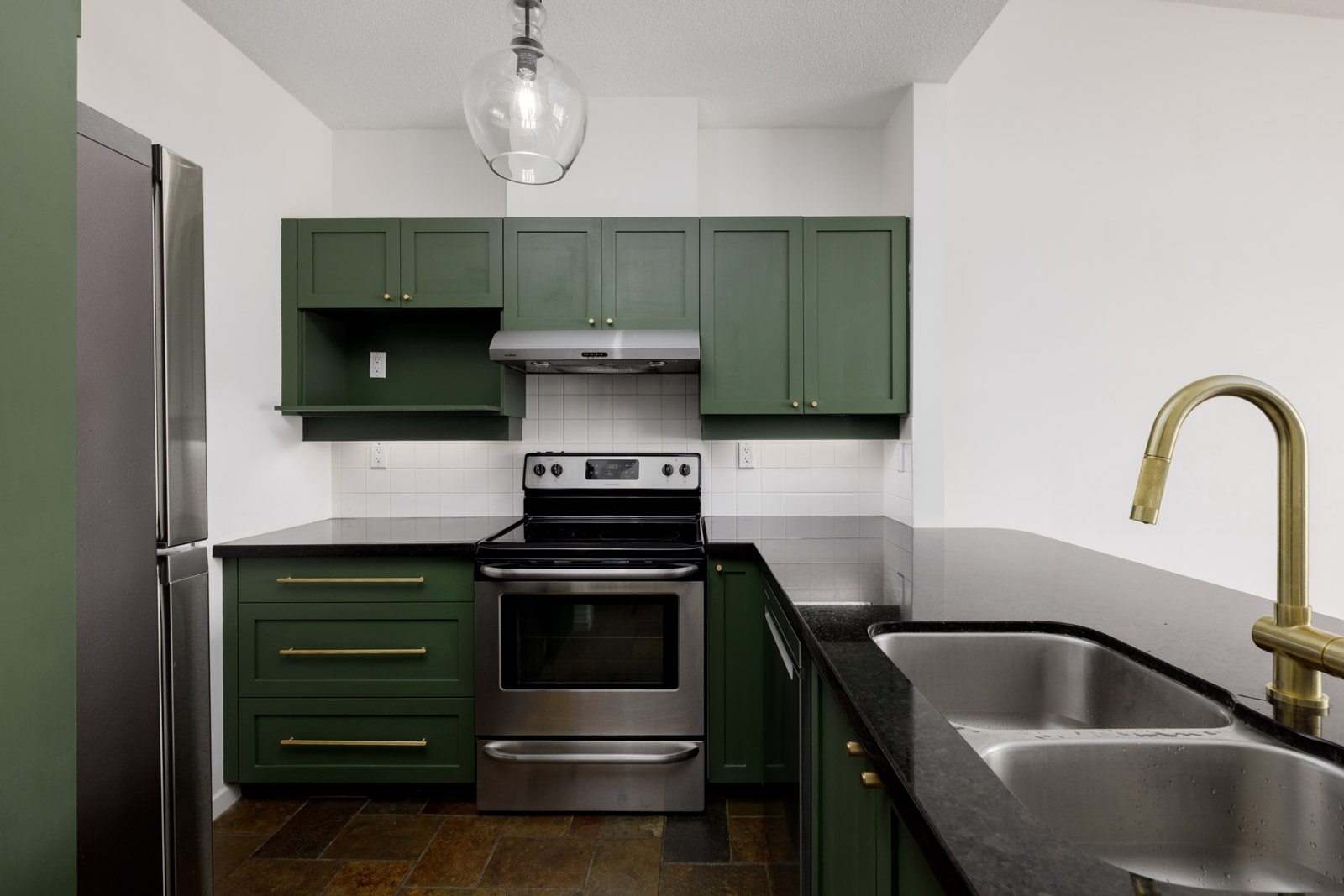 Modern kitchen with green cabinets, stainless steel appliances, black countertops, a double sink with a gold faucet, and white tile backsplash.