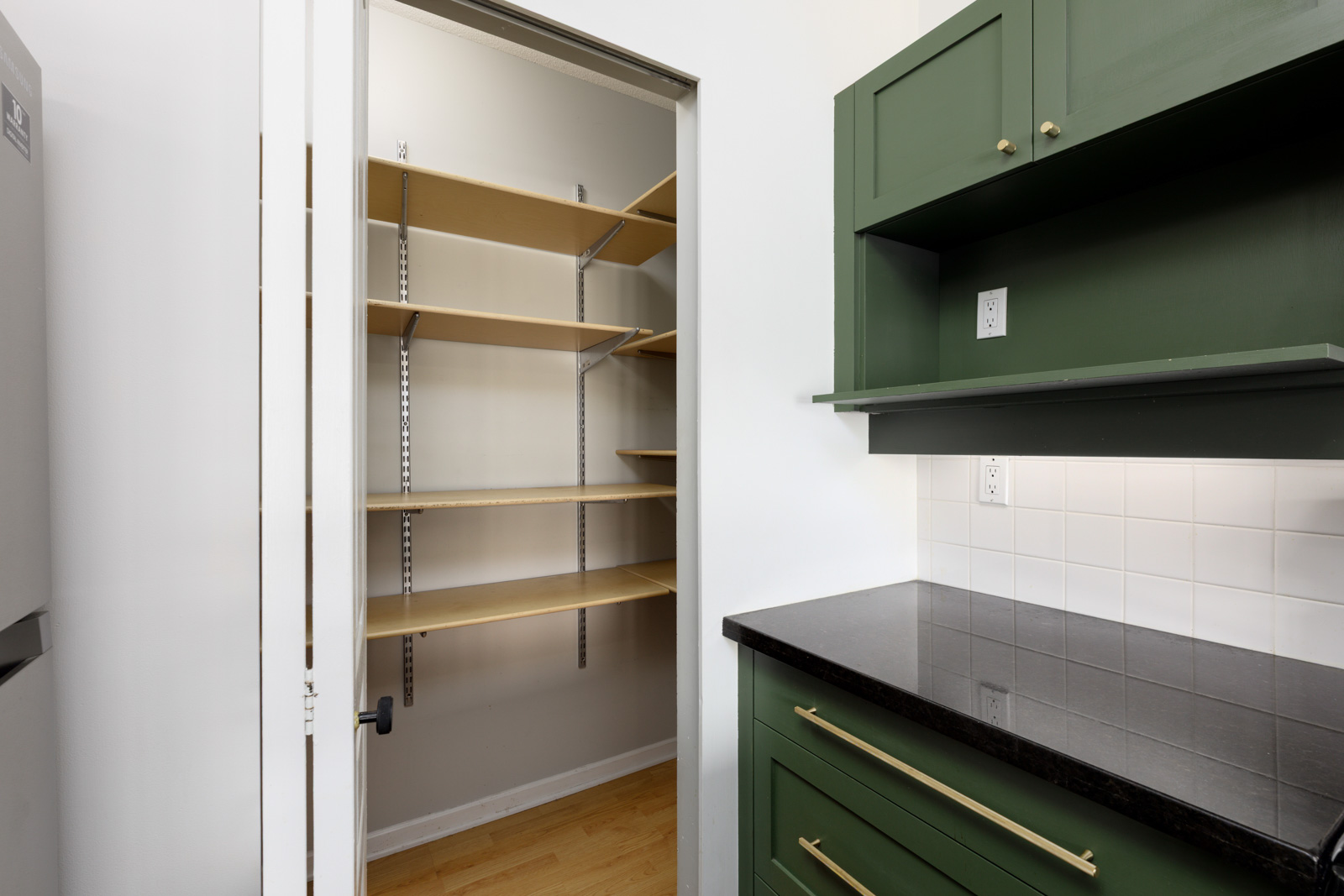 A kitchen with green cabinets, black countertop, white tile backsplash, and an open pantry door revealing empty wooden shelves.