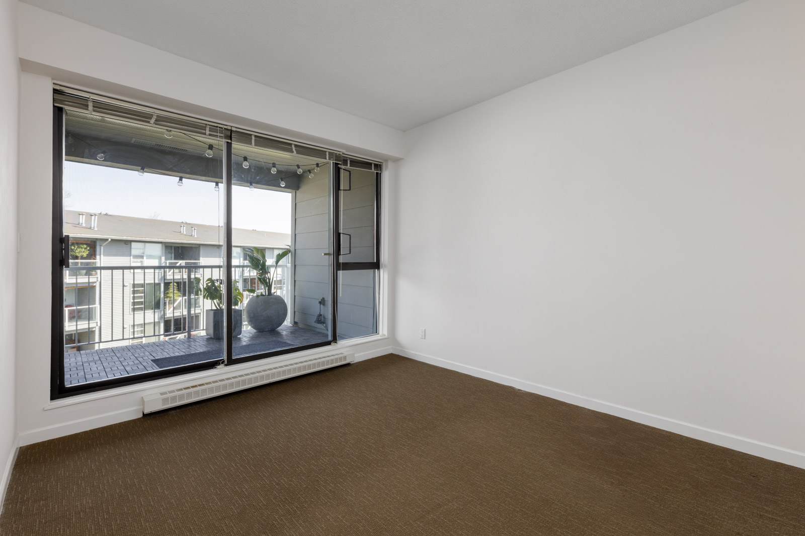 Empty room with beige carpet, white walls, and a large sliding glass door leading to a balcony with a potted plant and string lights.