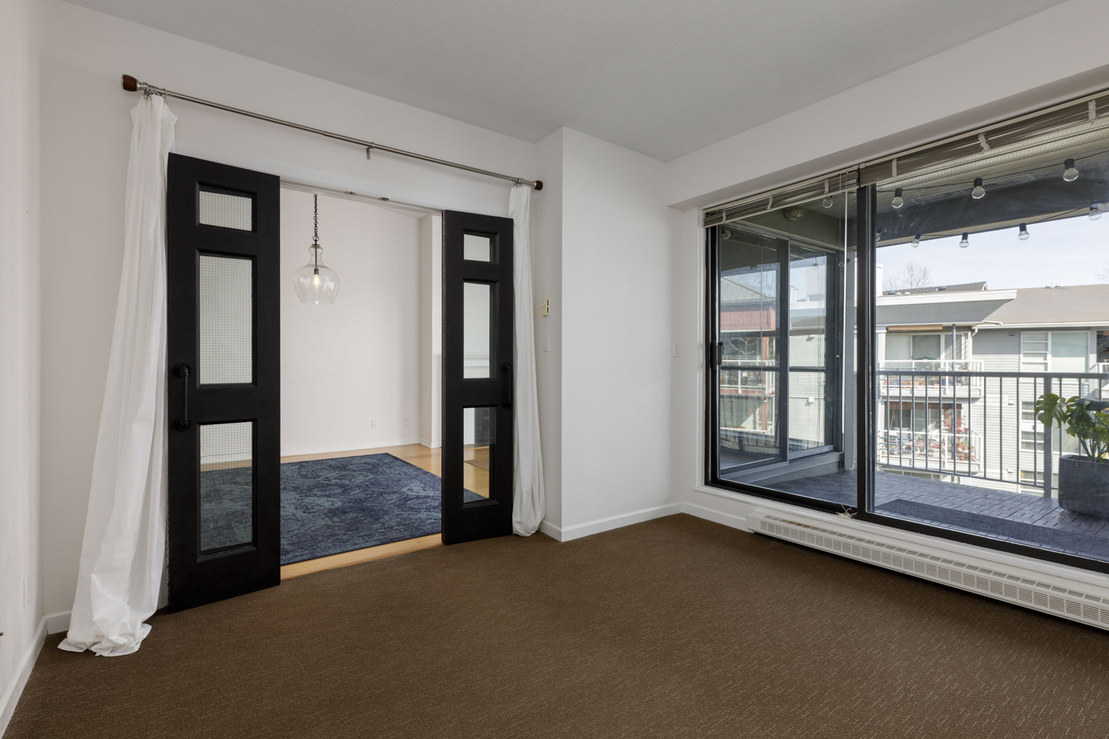Empty room with brown carpet, black-framed glass doors leading to another room, and large sliding glass doors opening to a balcony.