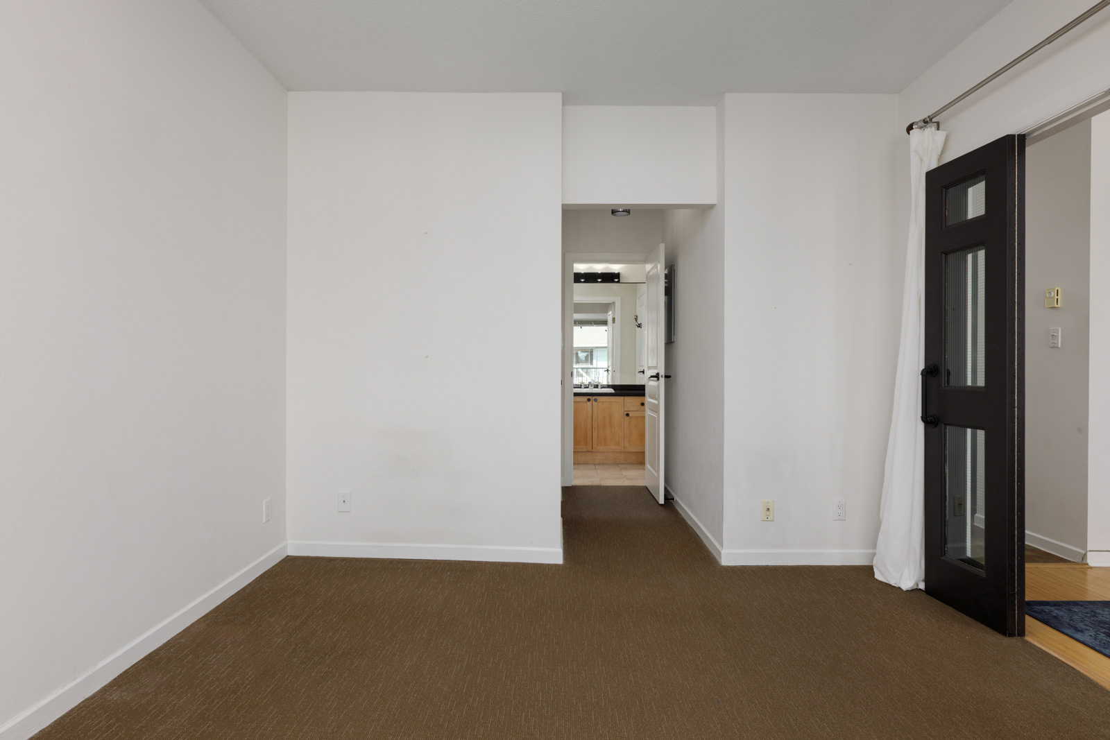Empty room with white walls, brown carpet, a doorway leading to a bathroom with a vanity, and a glass-panel door partially covered by a white curtain on the right.