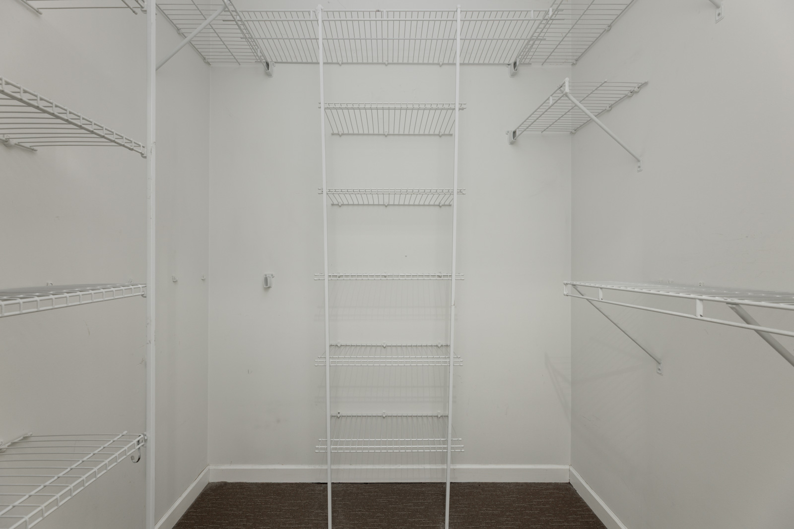 Empty walk-in closet with white wire shelving on the walls and a central shelving unit, white walls, and a dark carpeted floor.