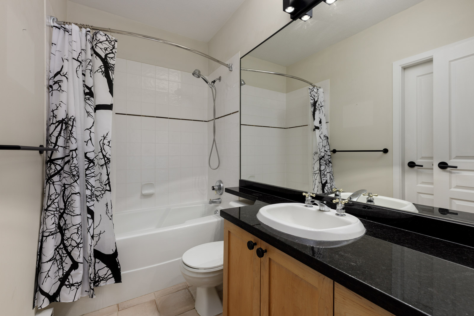 Modern bathroom with a bathtub and shower, white curtain with black tree design, black countertop, white sink, large mirror, and wooden cabinets.