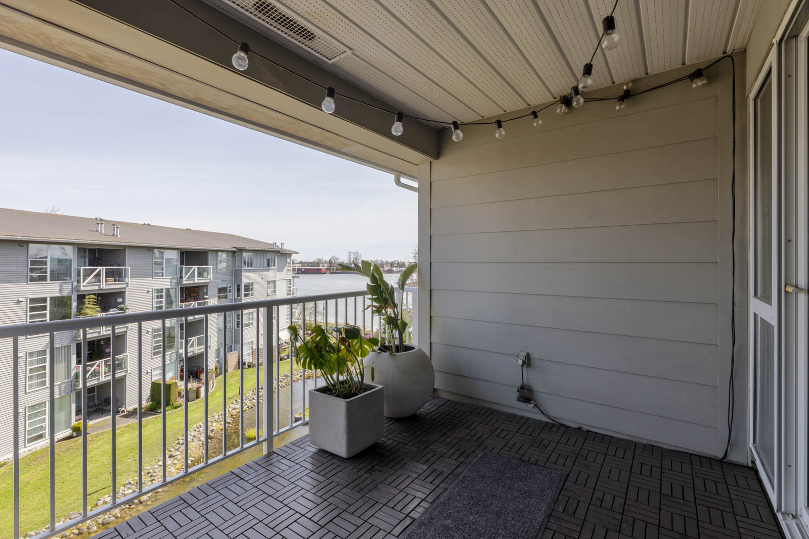 A covered balcony with string lights, two potted plants, and a view of nearby apartment buildings and water in the background.