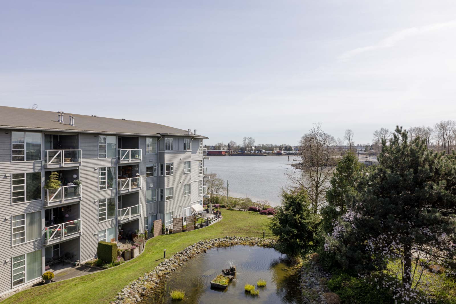A multi-story apartment building overlooks a landscaped pond and trees, with a river and distant buildings visible in the background under a clear sky.