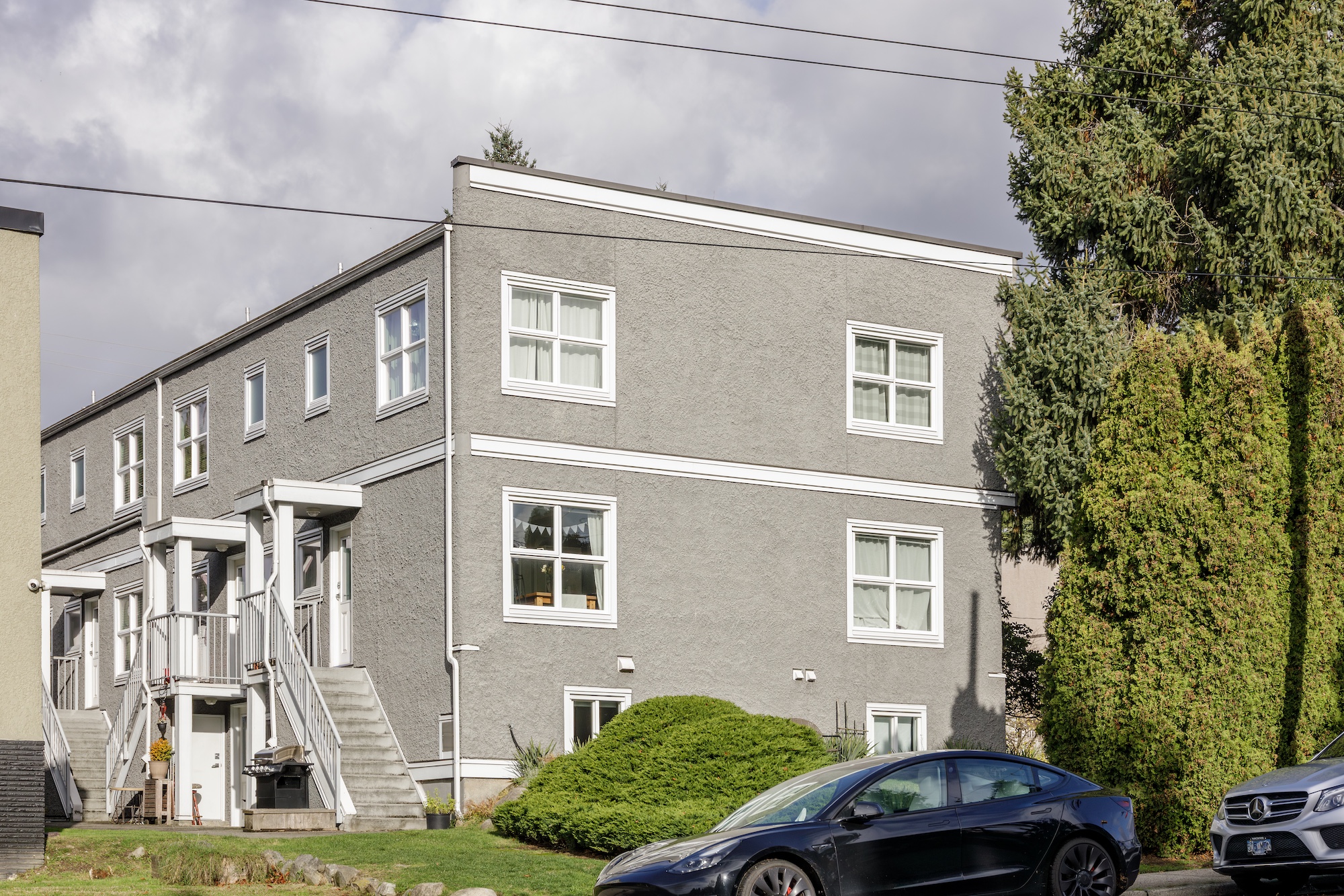 A two-story gray apartment building with white-trimmed windows, two exterior staircases, and cars parked in front on a cloudy day.