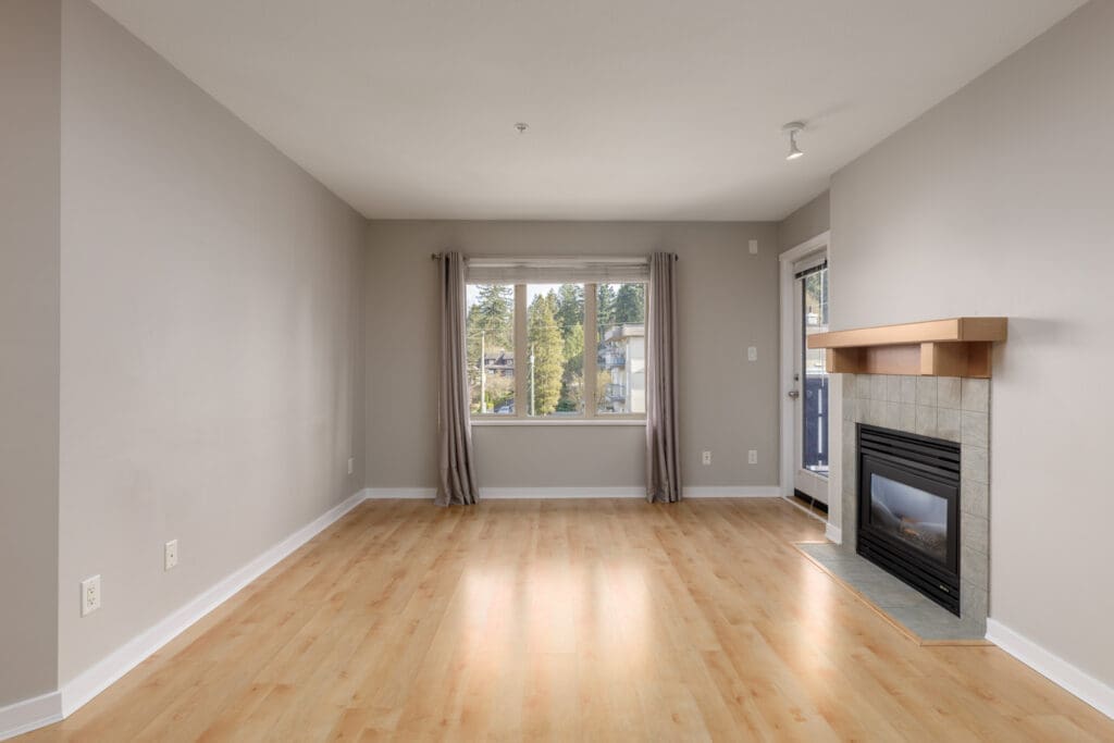 Empty living room with light wood flooring, beige walls, a window with curtains, and a corner fireplace with a wooden mantel.