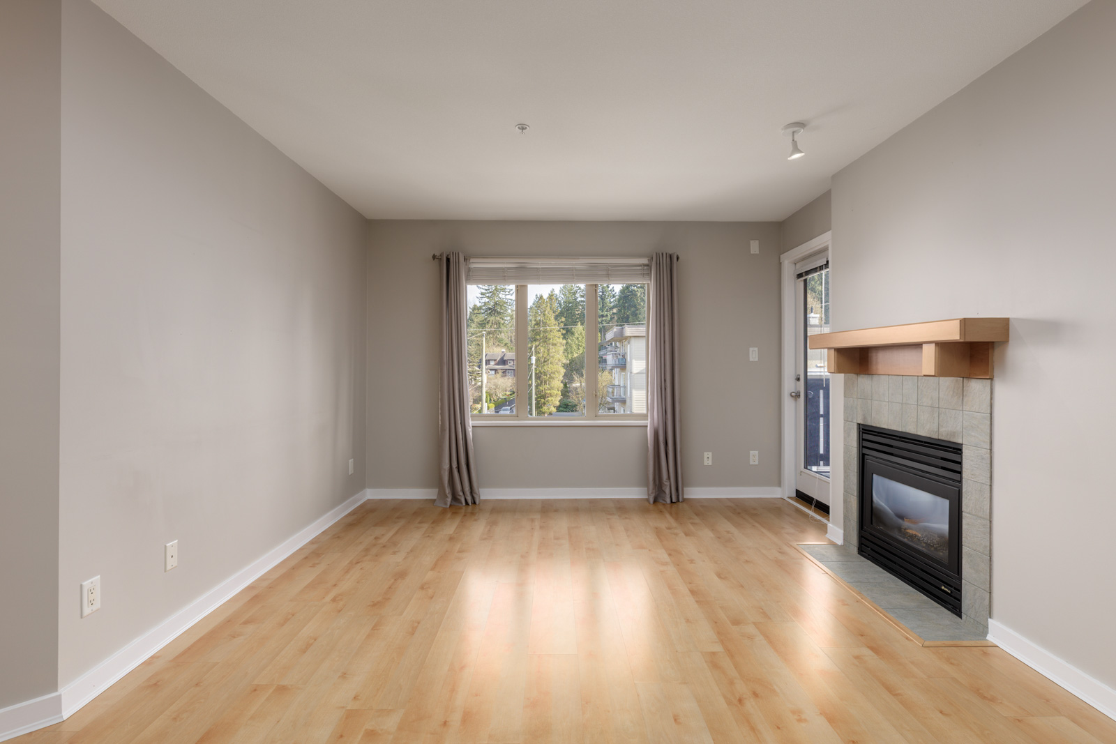 Empty living room with light wood flooring, beige walls, a window with curtains, and a corner fireplace with a wooden mantel.