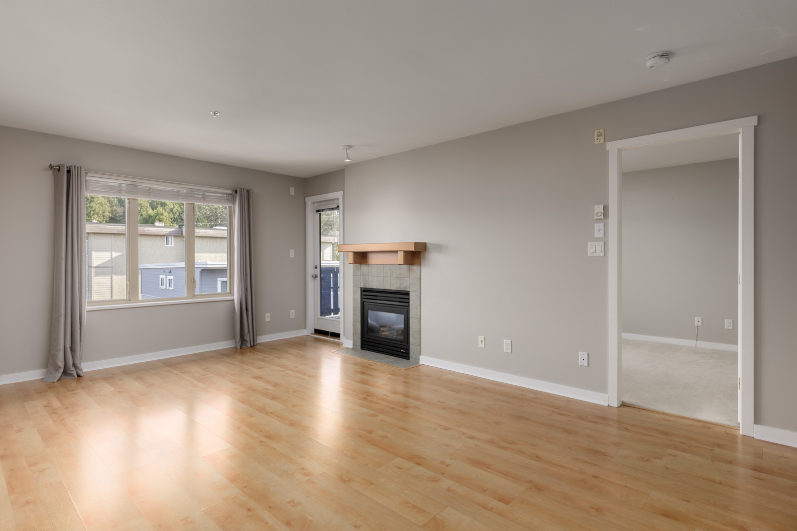 Unfurnished living room with light wood flooring, a fireplace, large window with gray curtains, and an open doorway leading to a carpeted room.