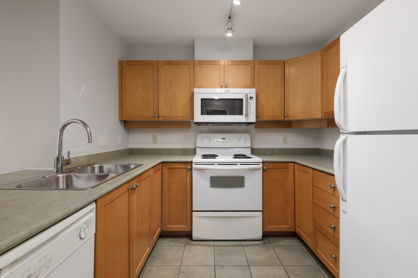 A kitchen with wooden cabinets, white appliances including a stove, microwave, refrigerator, dishwasher, and a stainless steel sink on a green countertop.