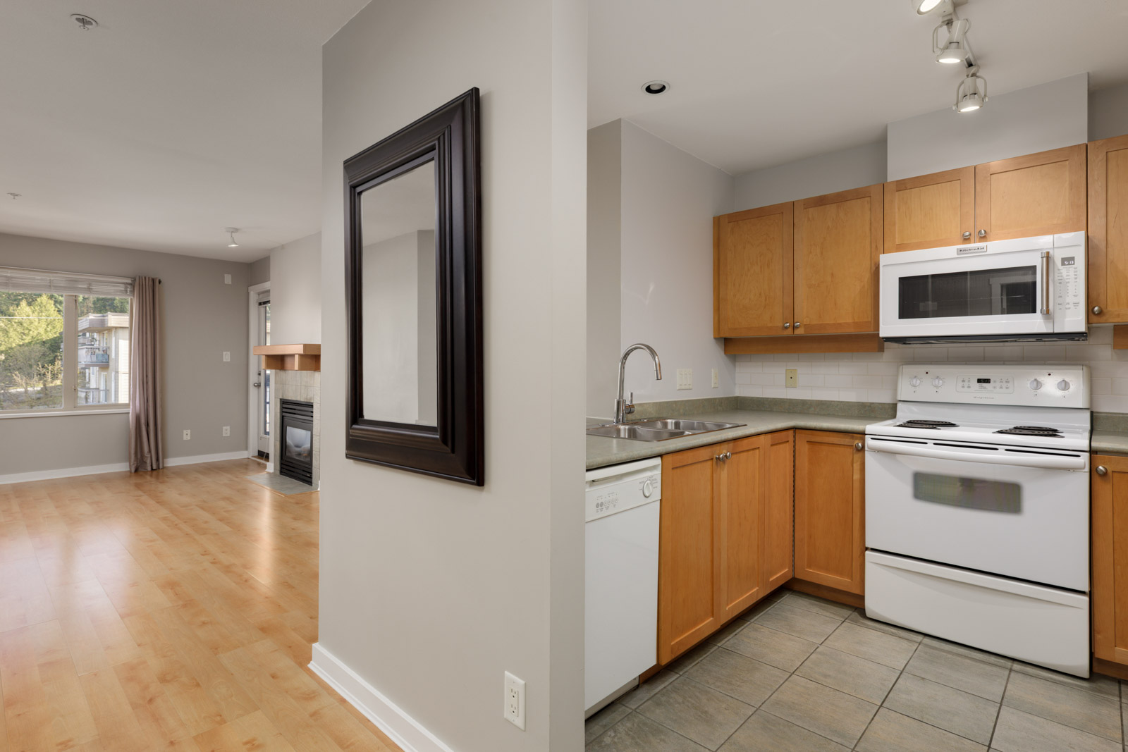 A modern apartment with a kitchen featuring wooden cabinets, white appliances, and tile flooring, adjacent to a living area with wood floors and a fireplace.