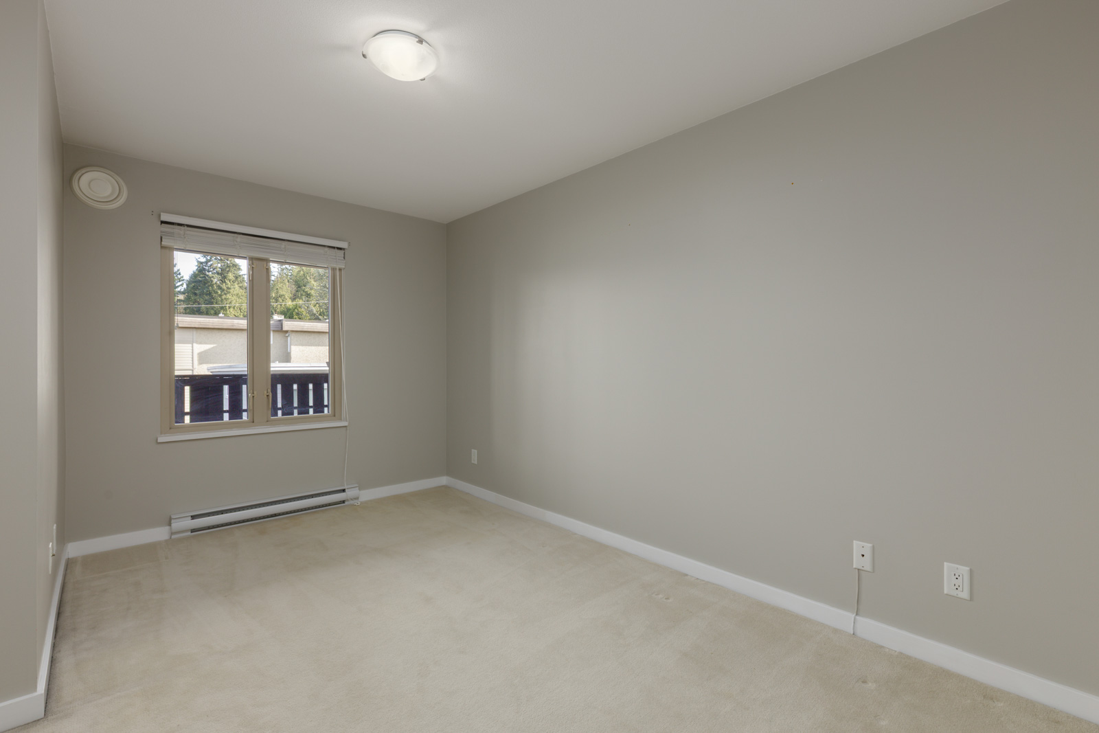 Empty room with beige carpet, light gray walls, a window with a view outside, and a ceiling light fixture. Electrical outlets are visible on the right wall.