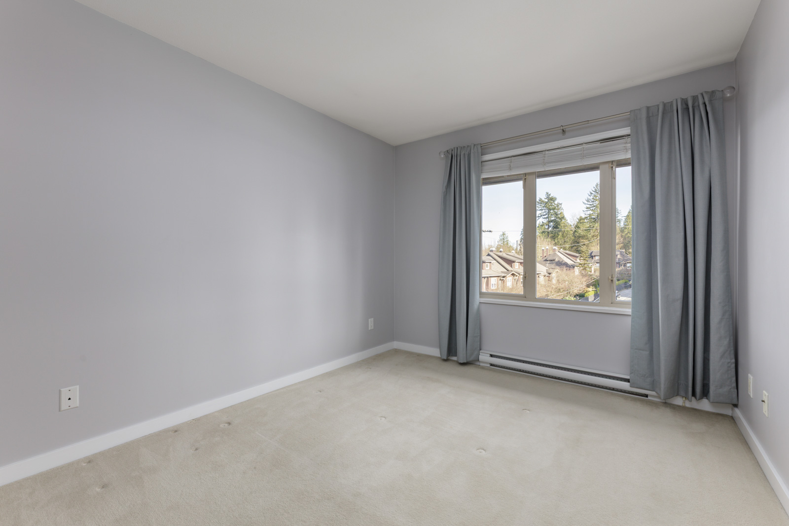 Empty bedroom with light gray walls, beige carpet, and a window with blue-gray curtains overlooking a residential neighborhood.
