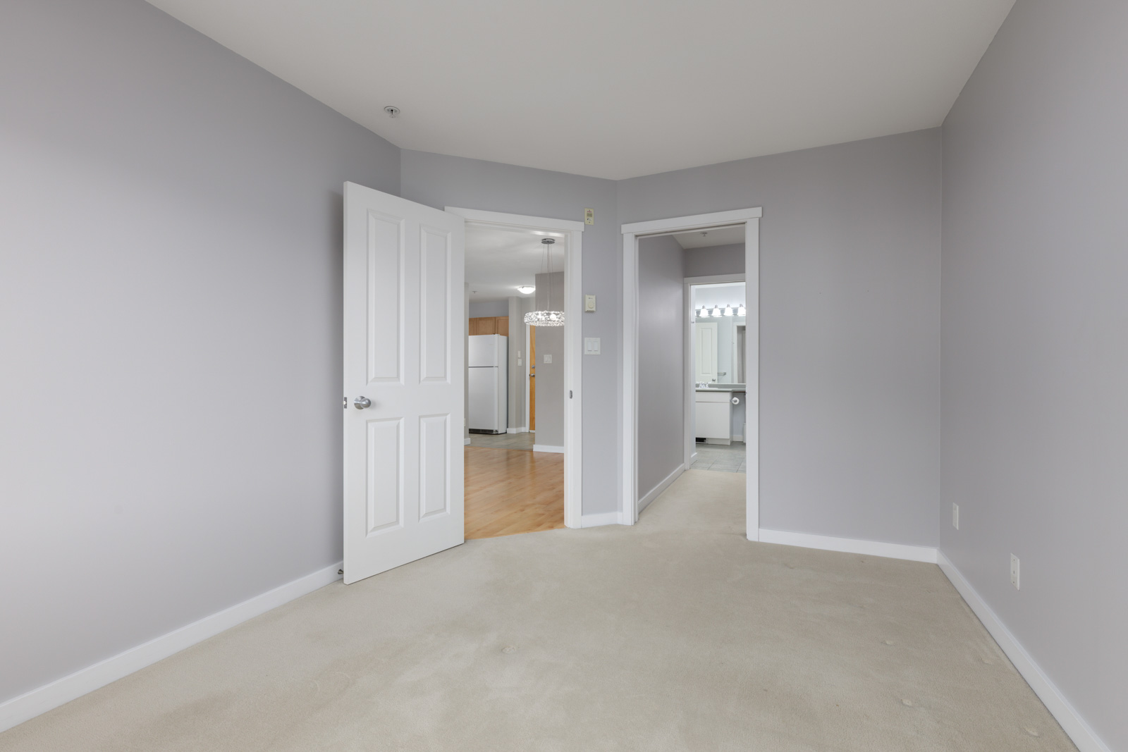 Empty bedroom with light gray walls and beige carpet, featuring two open doors leading to a kitchen area and a bathroom.