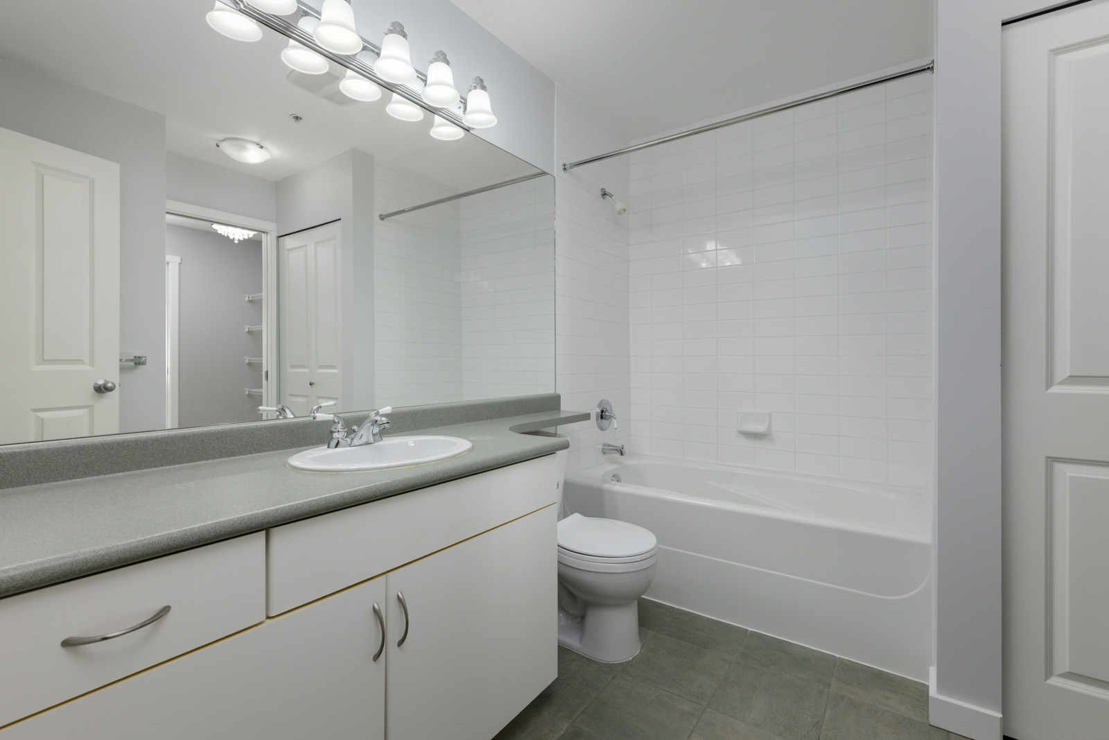 Modern bathroom with a large mirror, white cabinets, gray countertop, toilet, and a bathtub with a white tiled wall. Bright overhead lighting.