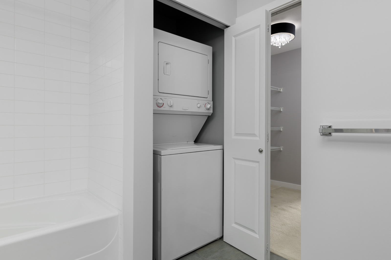 A stacked washer and dryer unit is installed in a closet with open double doors, adjacent to a white tiled bathtub in a modern bathroom.