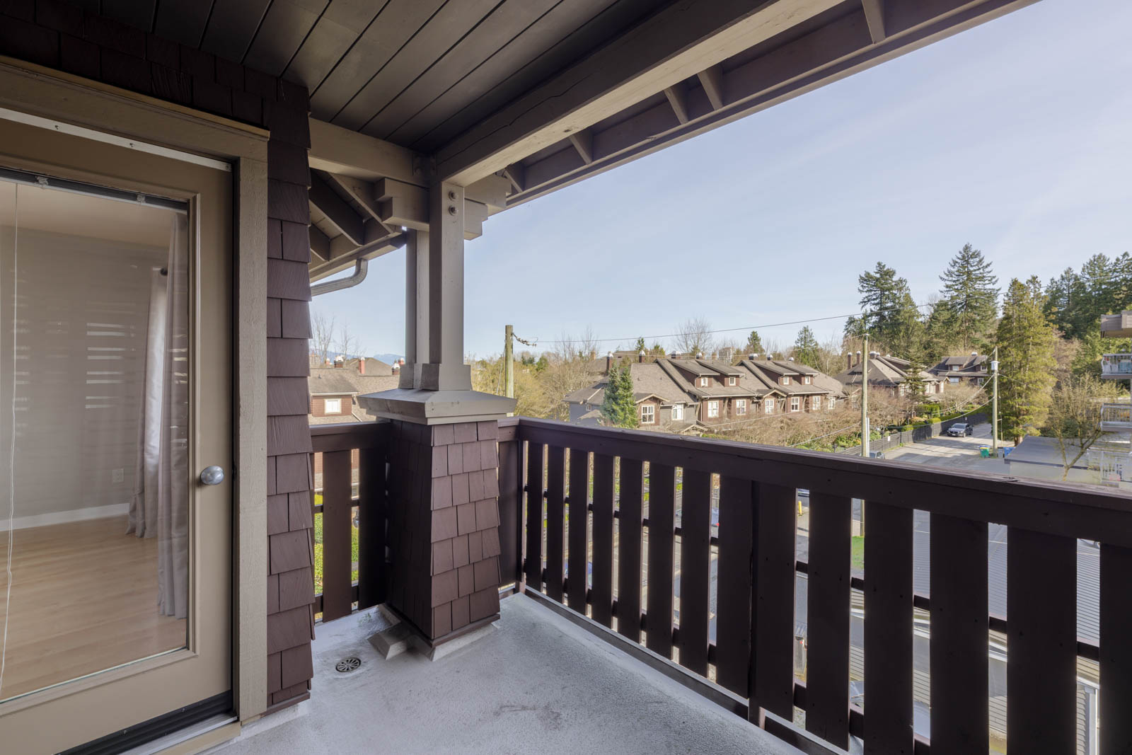 View from a covered balcony with brown wooden railing, looking out over a suburban neighborhood with houses, trees, and a clear sky.