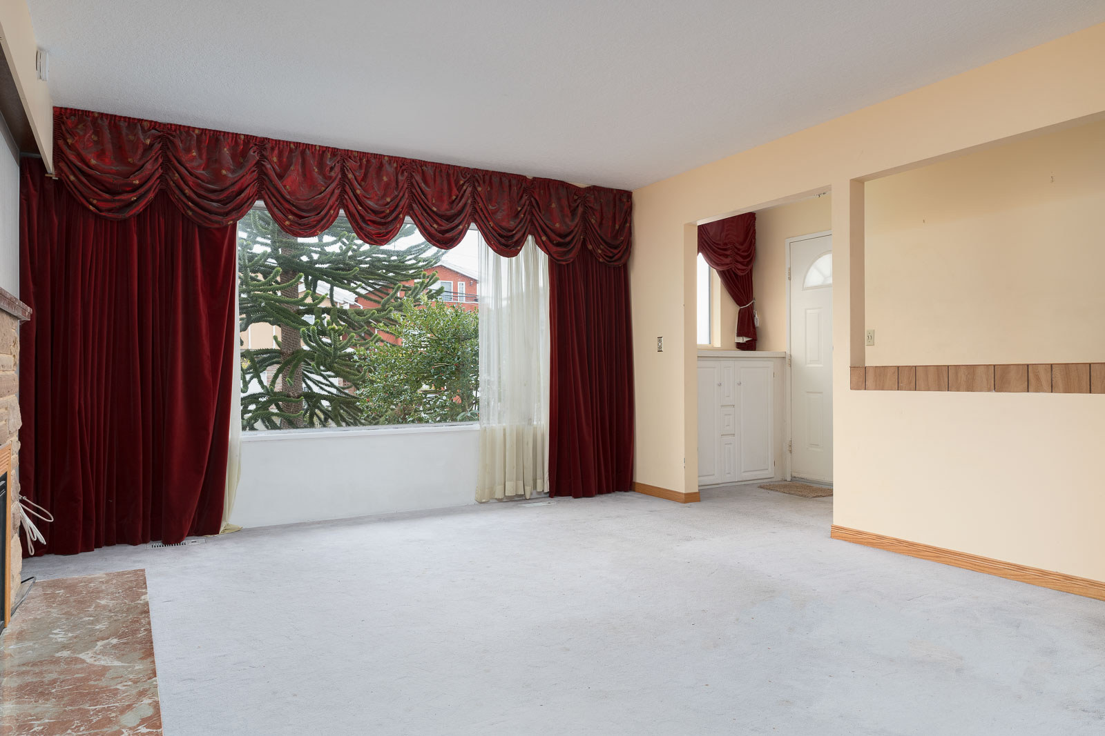 Empty living room with red curtains, a large window showing greenery outside, off-white walls, and an entryway with a small step.