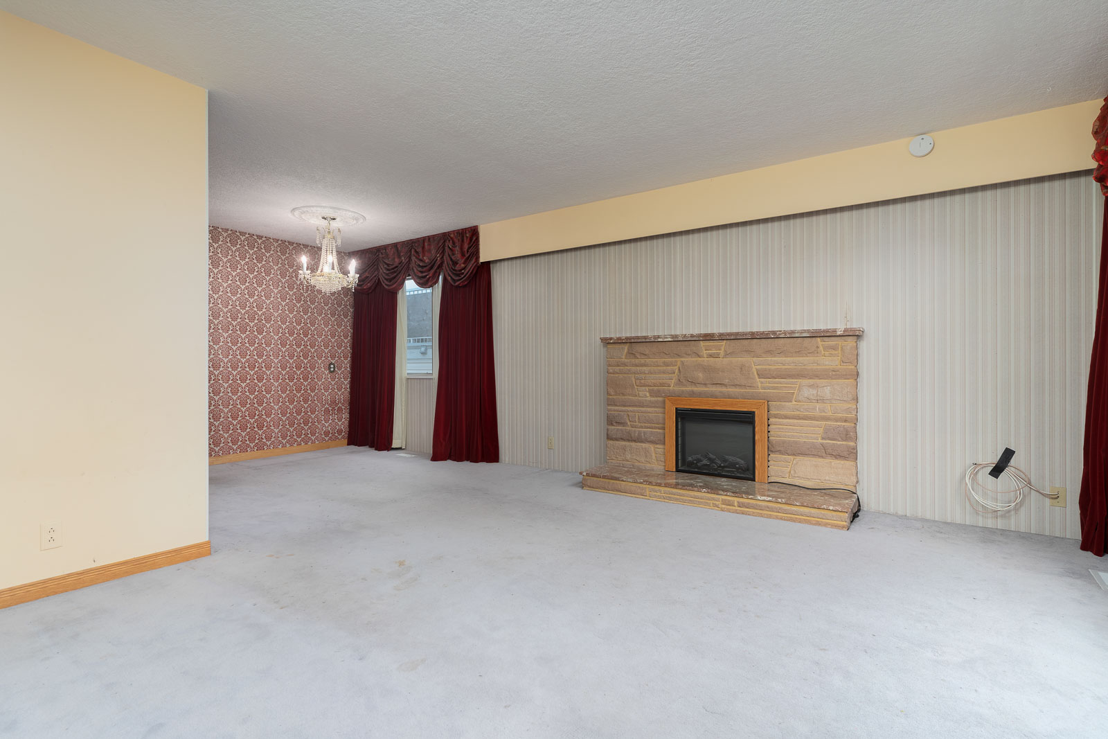 Living room with stone fireplace, light carpet, large window with vertical blinds and red curtains, adjacent dining area with chandelier, and patterned wallpaper.