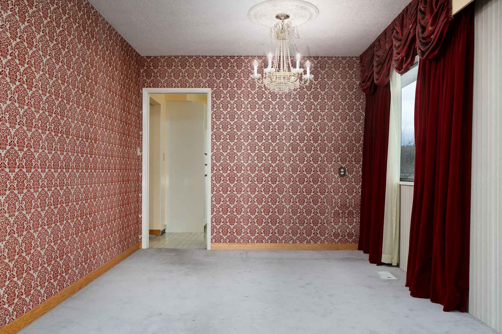 An empty room with patterned red wallpaper, a chandelier, red curtains, a window, and an open doorway leading to a hallway.