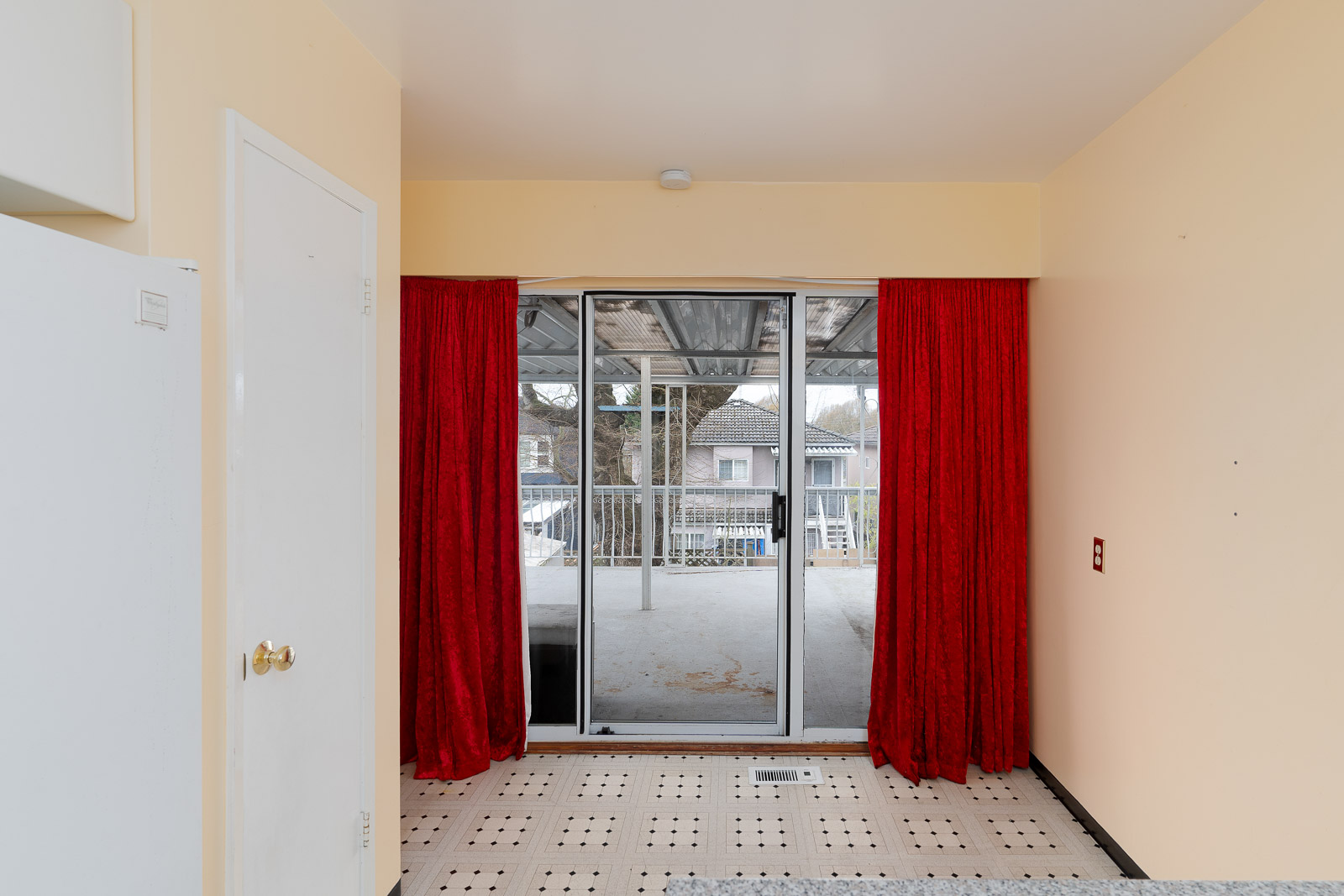 Sliding glass door with red curtains opens to a covered patio; view of neighboring houses visible outside. Tiled floor and cream-colored walls inside.