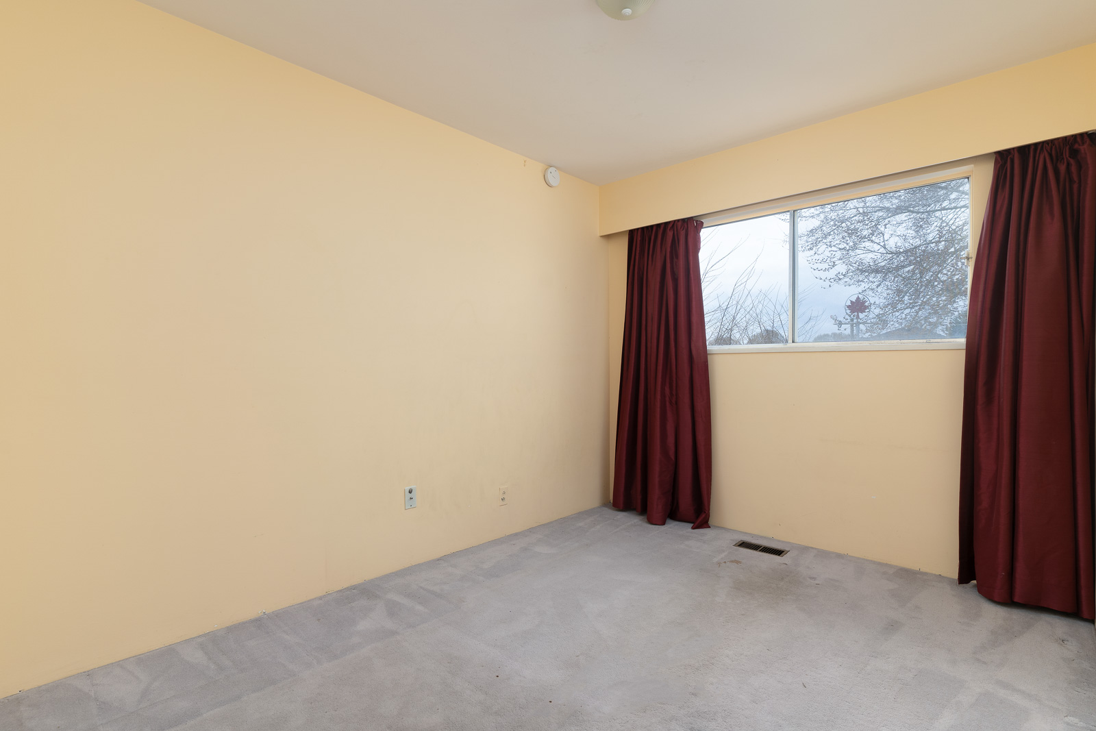 Empty room with beige walls, light gray carpet, a large window with red curtains, and visible power outlets on one wall.