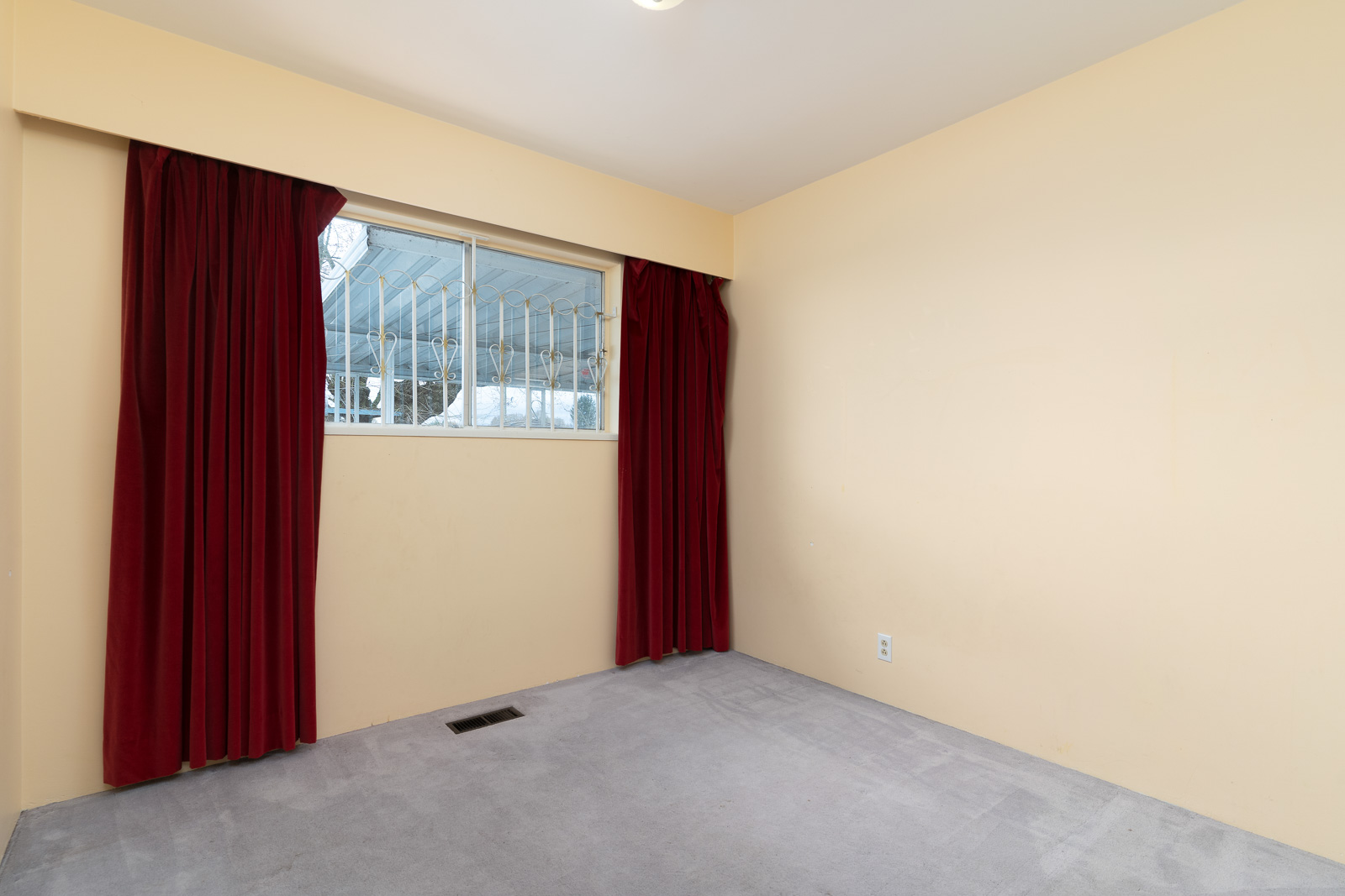 Empty room with beige walls, gray carpet, a window with security bars, and closed red curtains.