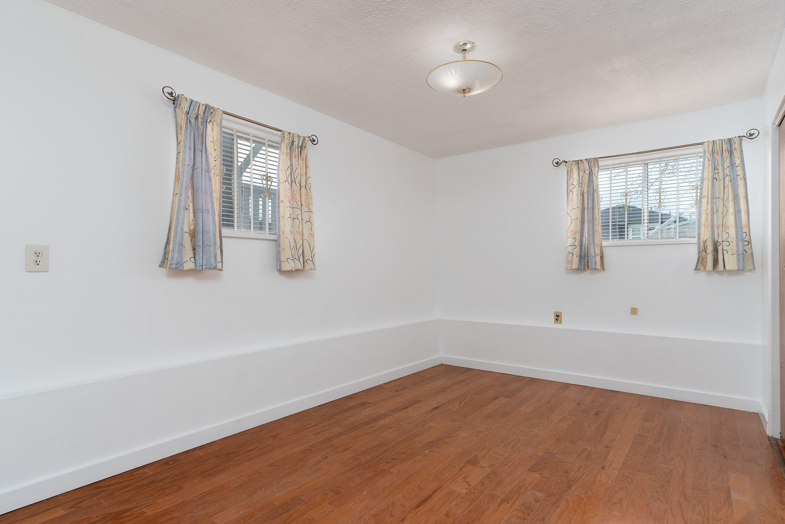 Unfurnished room with hardwood floor, white walls, two windows with patterned curtains, and a ceiling light fixture.