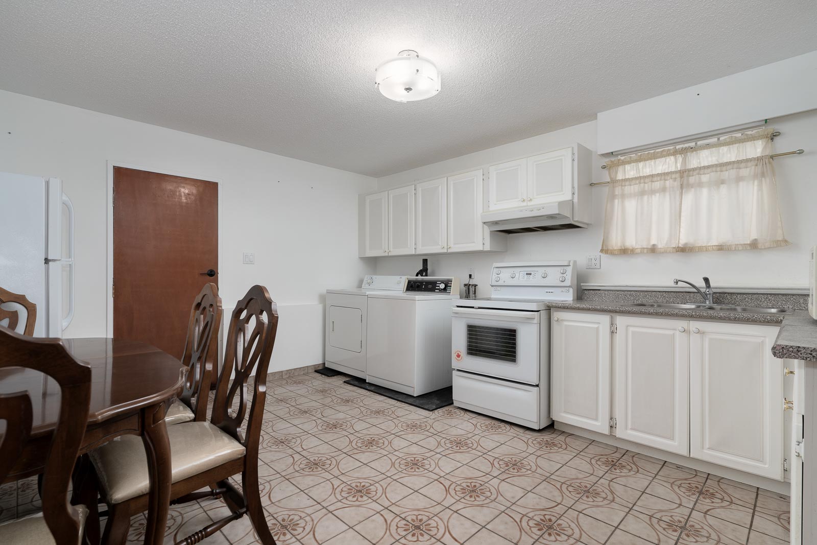 Kitchen with white cabinets, white appliances, a dining table with chairs, tiled floor, and a window with sheer curtains.