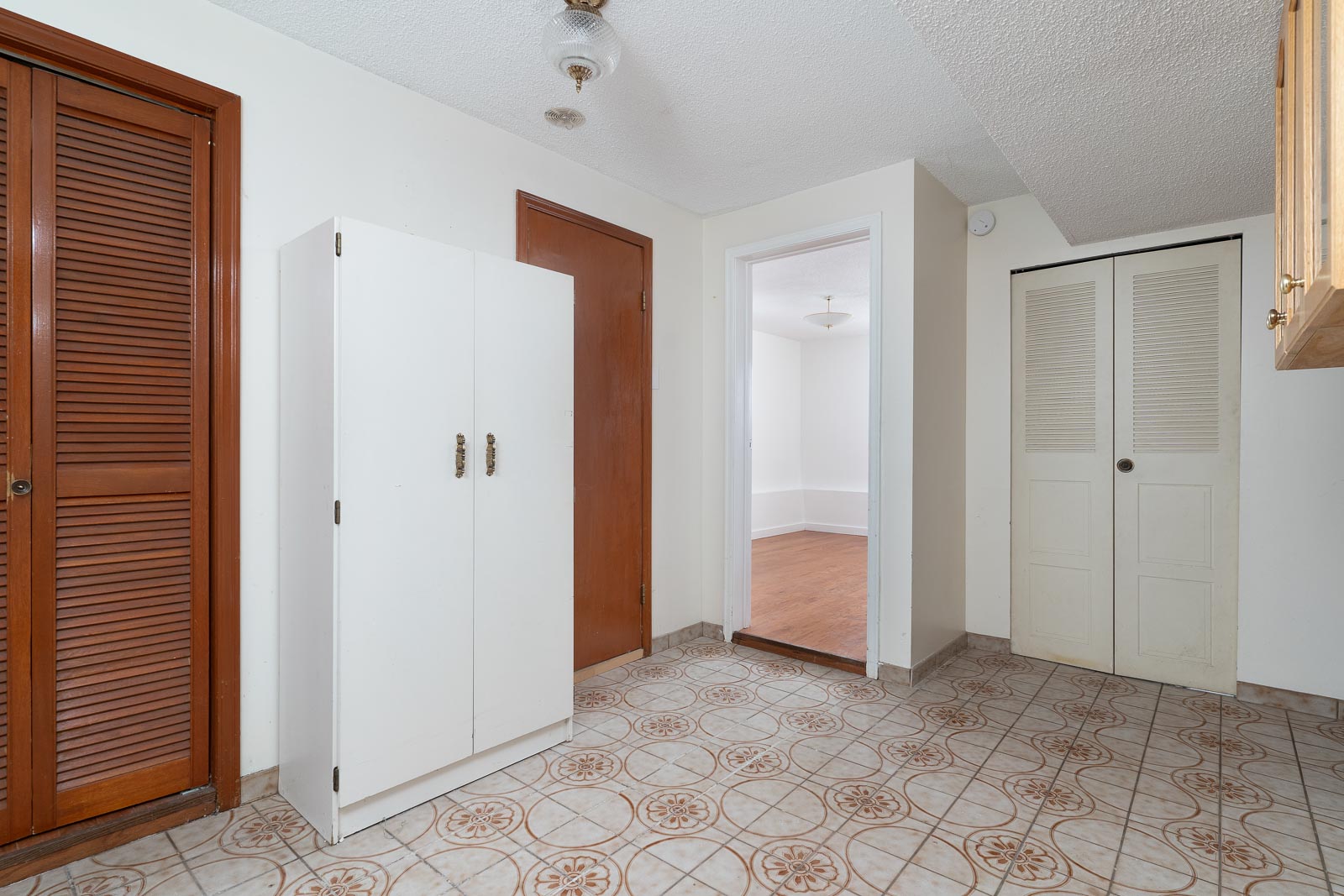 Room with patterned tile floor, white and wood doors, a white cabinet, and view into an adjacent room with wood flooring and a ceiling light.