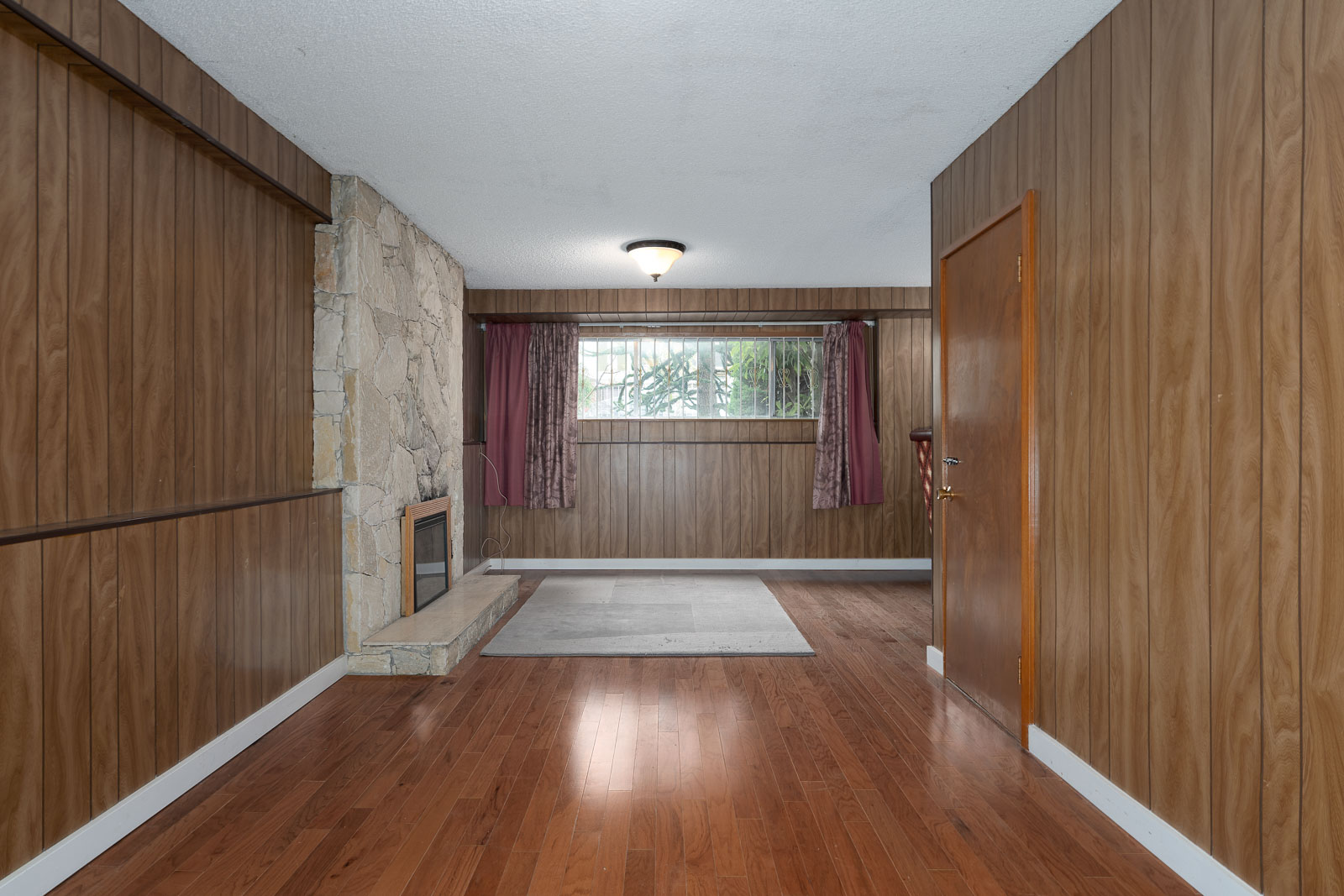 Room with wood-paneled walls, hardwood floor, stone fireplace, window with purple curtains, and a ceiling light fixture.