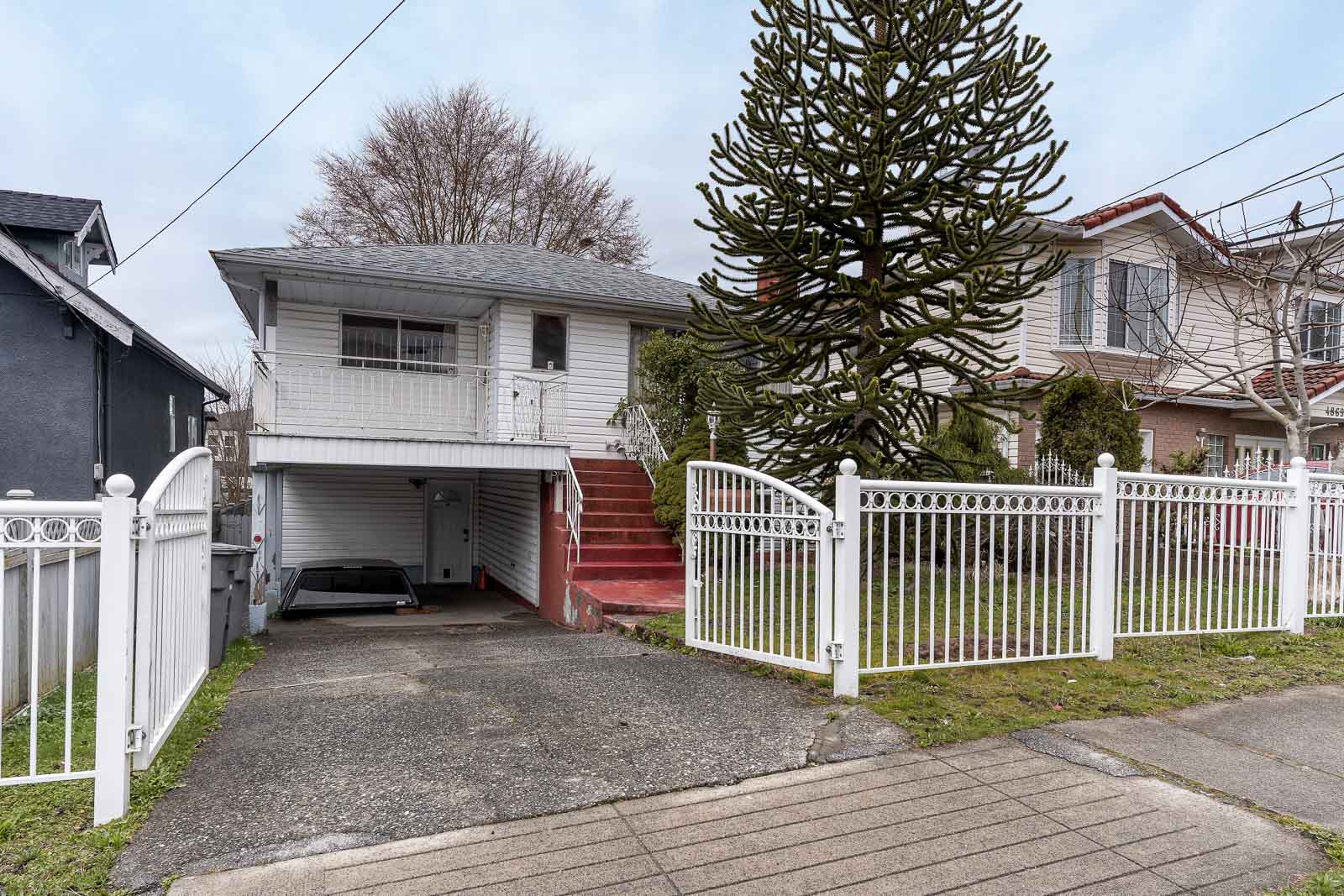 A two-story white house with a red staircase, metal fence, driveway, and large tree in the front yard, situated in a residential neighborhood.