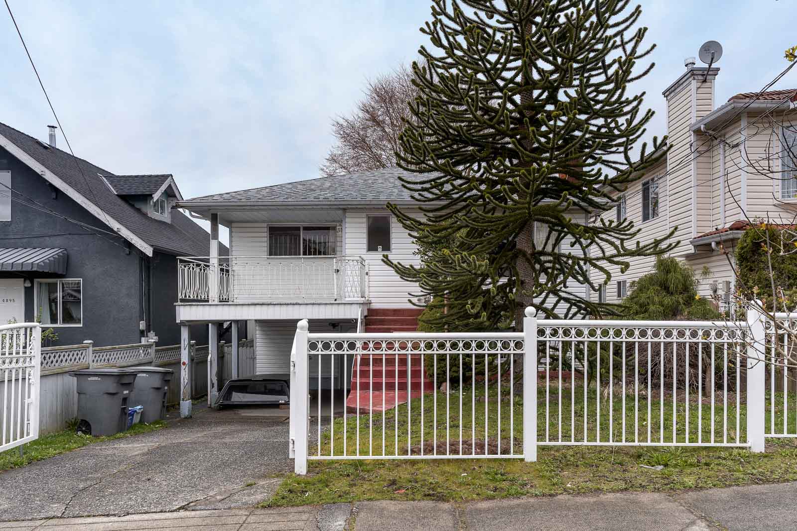 A two-story white house with a balcony, white fence, large tree in the front yard, and a paved driveway leading to a carport.