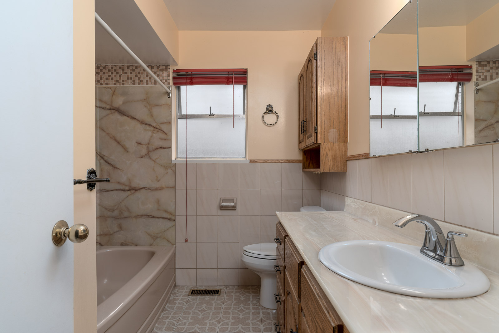 Bathroom with a bathtub, toilet, and sink. Wooden cabinets and drawers, large mirror, two frosted windows, and beige tile on walls and floor.