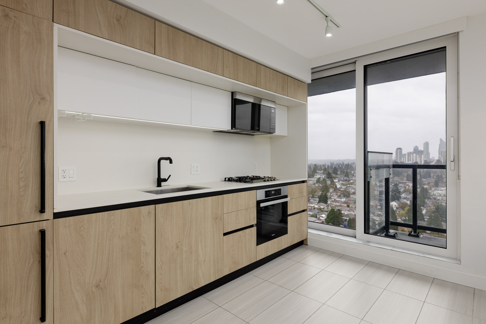 Modern kitchen with light wood cabinets, built-in appliances, and a large window leading to a balcony with a city view.