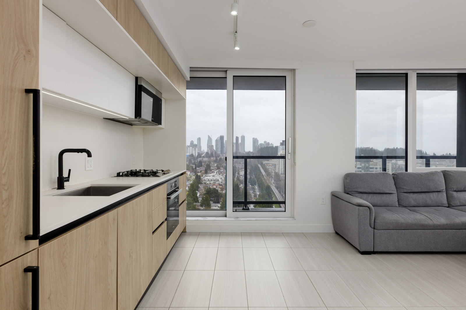 Modern apartment kitchen with light wood cabinets, built-in appliances, and a gray sofa near large windows overlooking a cityscape on a cloudy day.