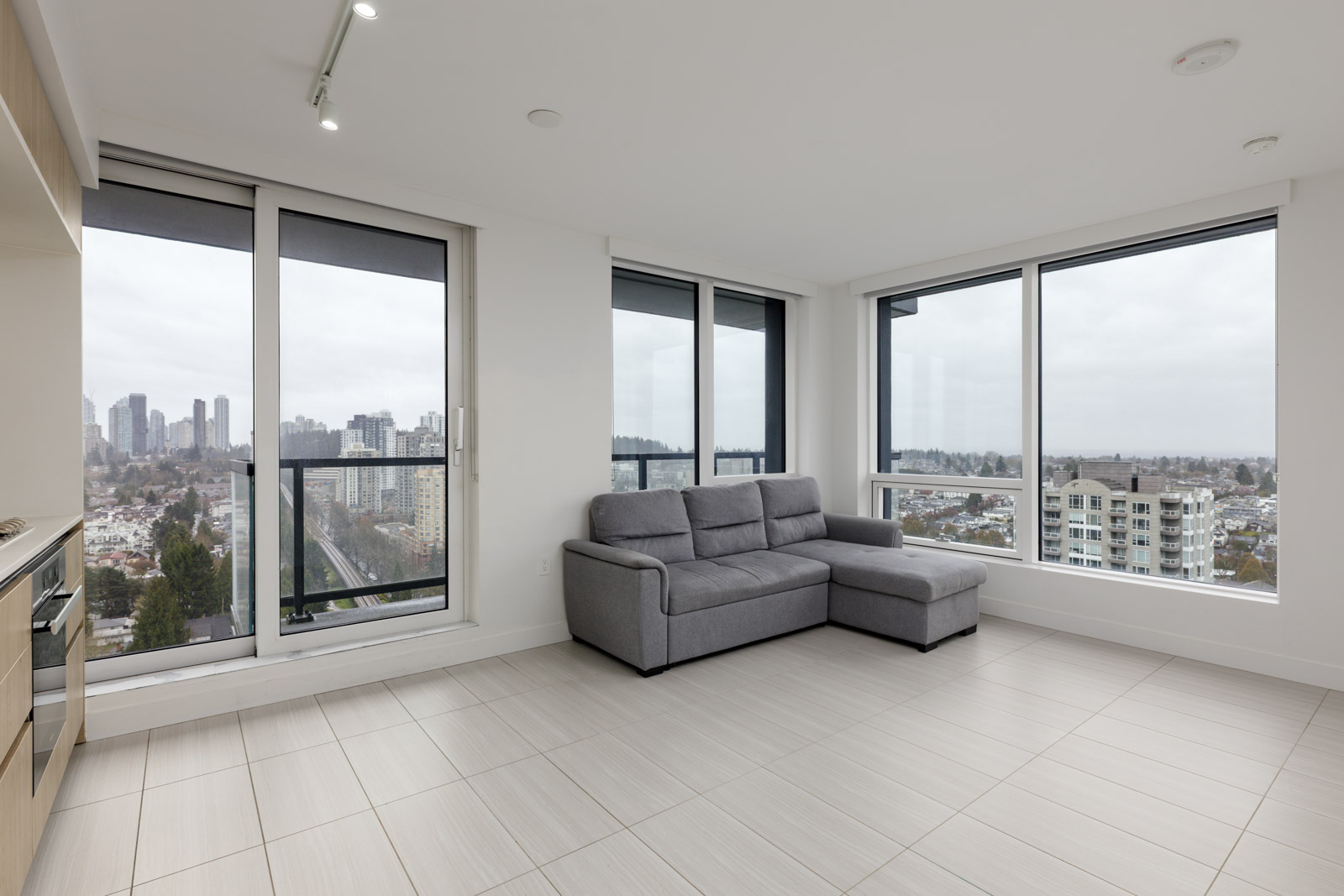 Modern, unfurnished living room with large windows, a gray sectional sofa, tiled floor, and cityscape views outside on a cloudy day.