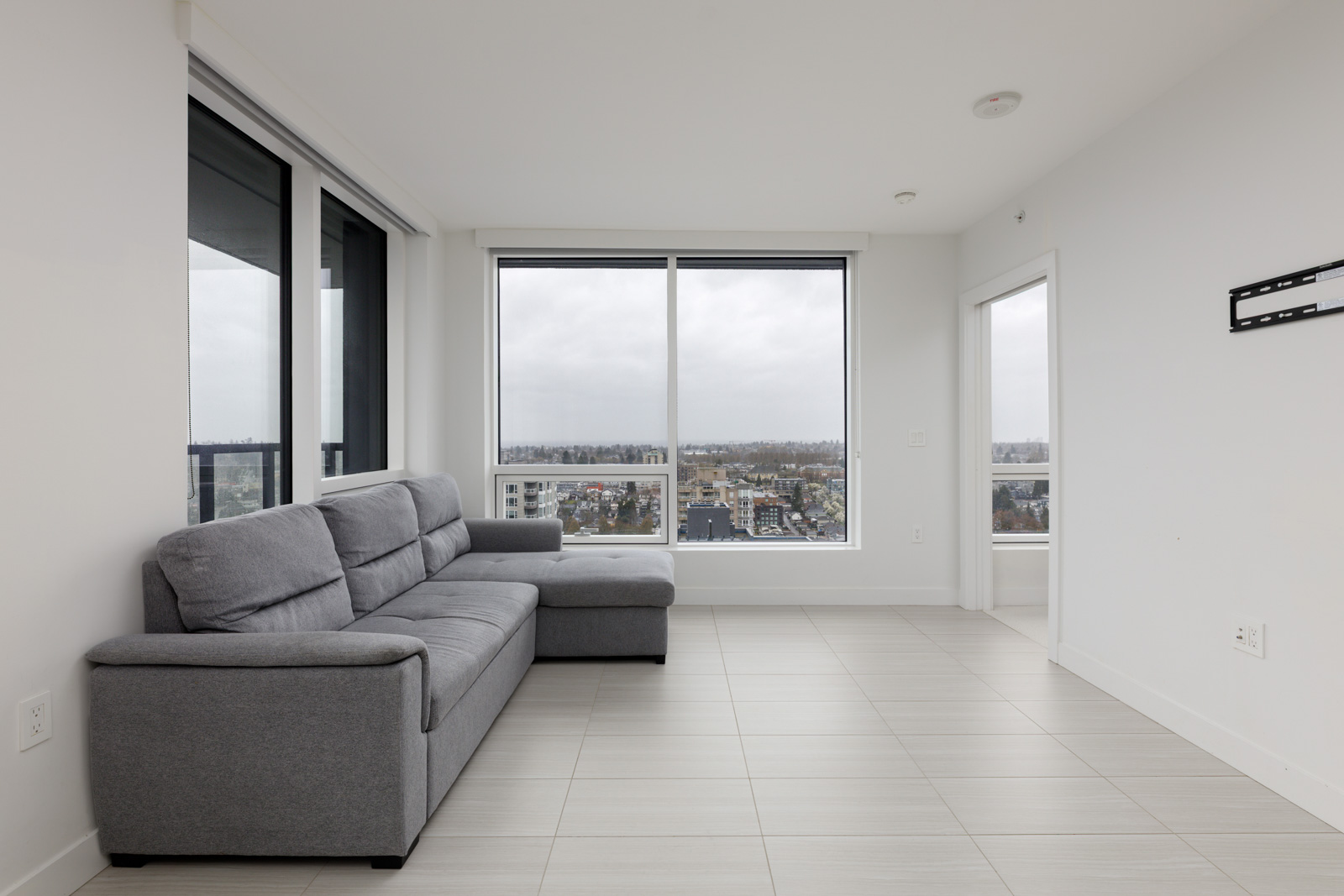 A modern, unfurnished living room with a gray sectional sofa, large windows overlooking a cityscape, and light tile flooring.