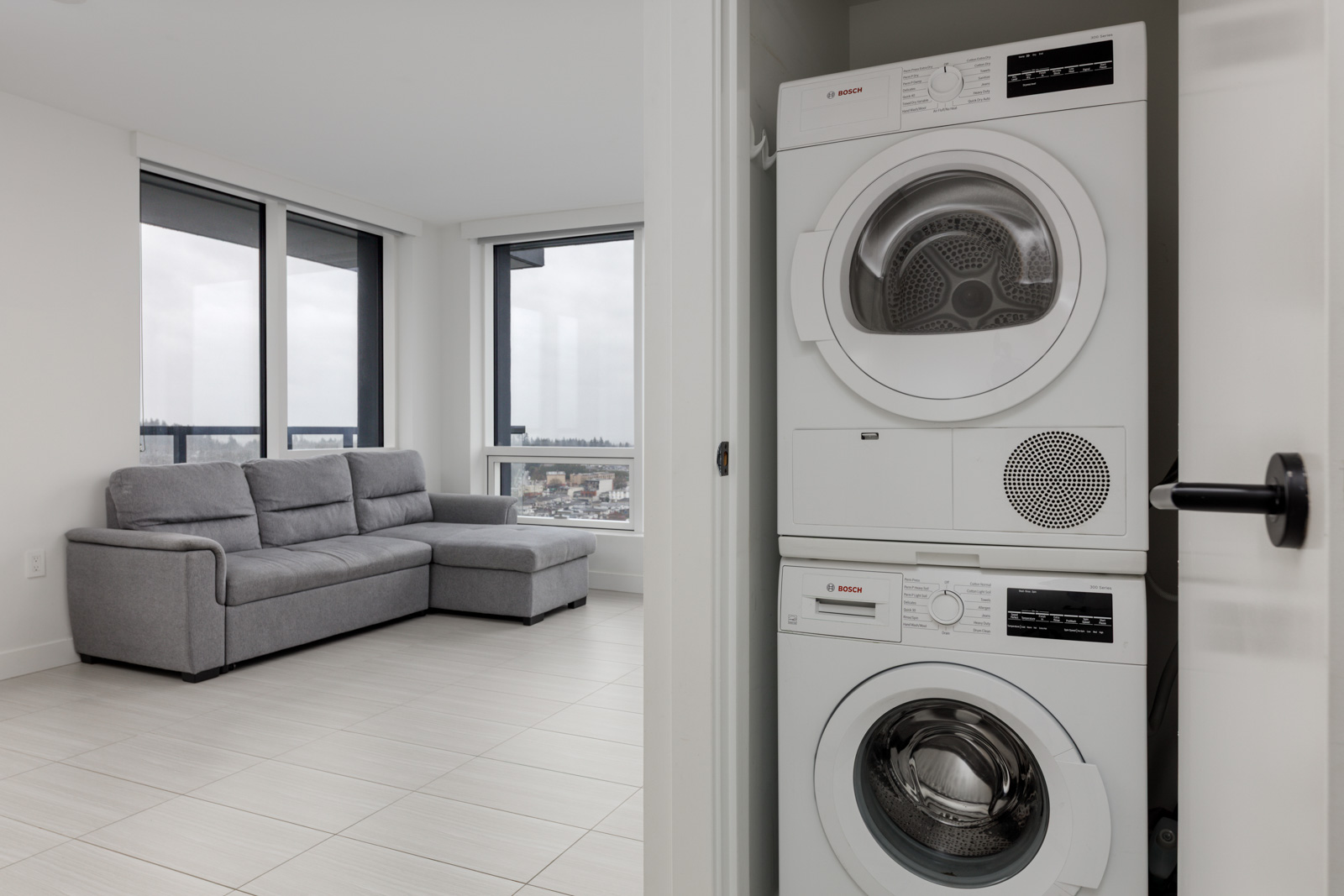A modern apartment with a gray sofa in a living area and a stacked Bosch washer and dryer unit in a closet. Large windows provide a view of the city.