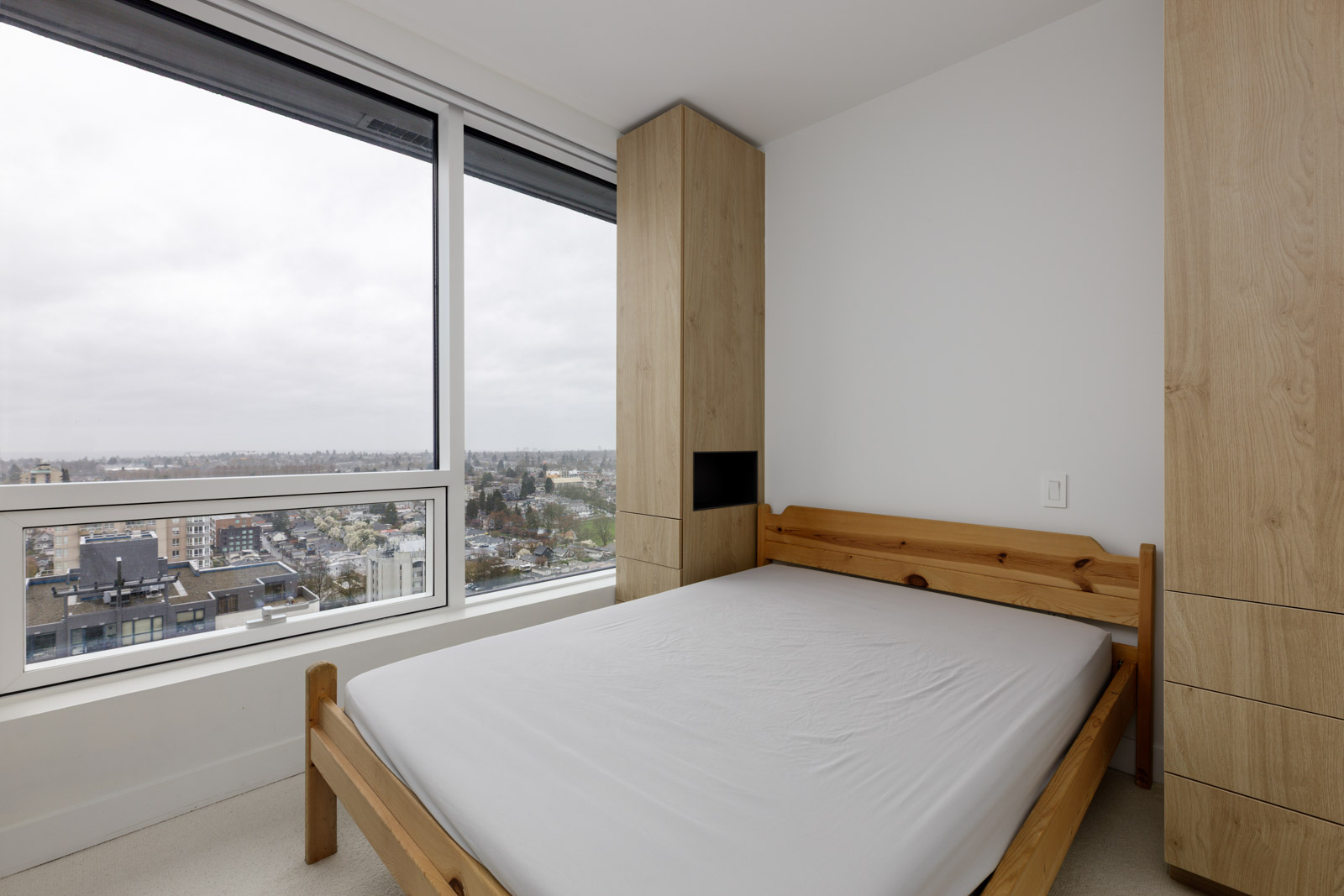 A small, modern bedroom with a wooden bed frame, built-in wooden wardrobes, large window, and a cityscape view on a cloudy day.