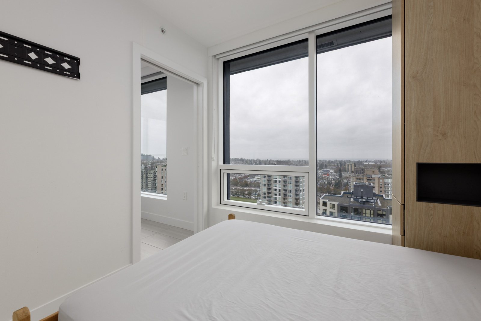 A modern, minimal bedroom with a large window showing a cityscape view on a cloudy day. A bed with white linens is in the foreground.