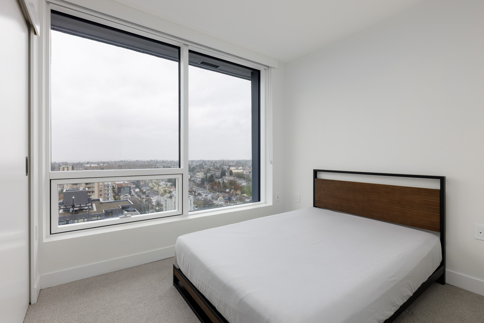 A minimalist bedroom with a bed against a white wall and large windows offering a cityscape view on an overcast day.