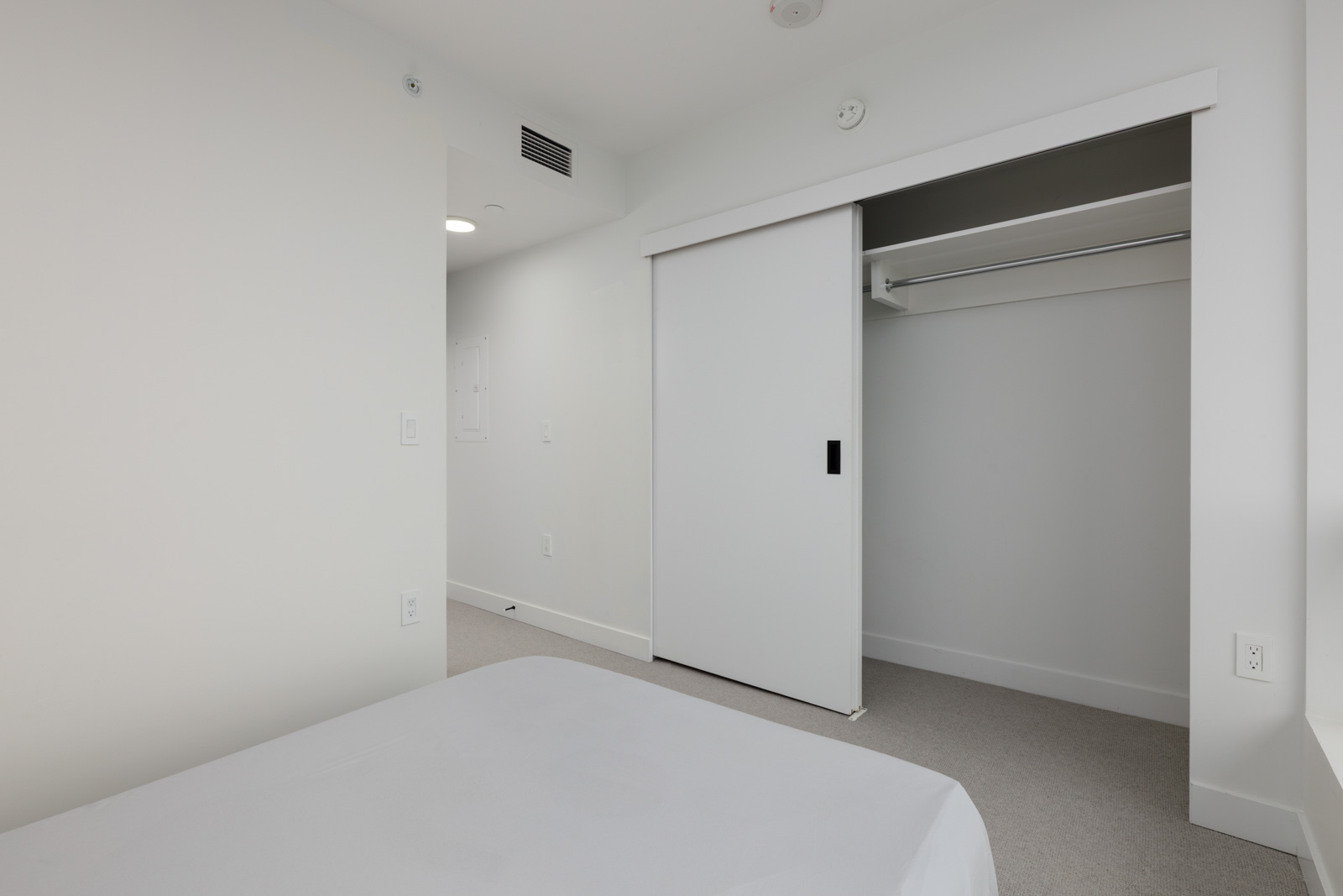 Minimalist bedroom with white walls, a bed with plain bedding in the foreground, and an open closet with sliding doors in the background.