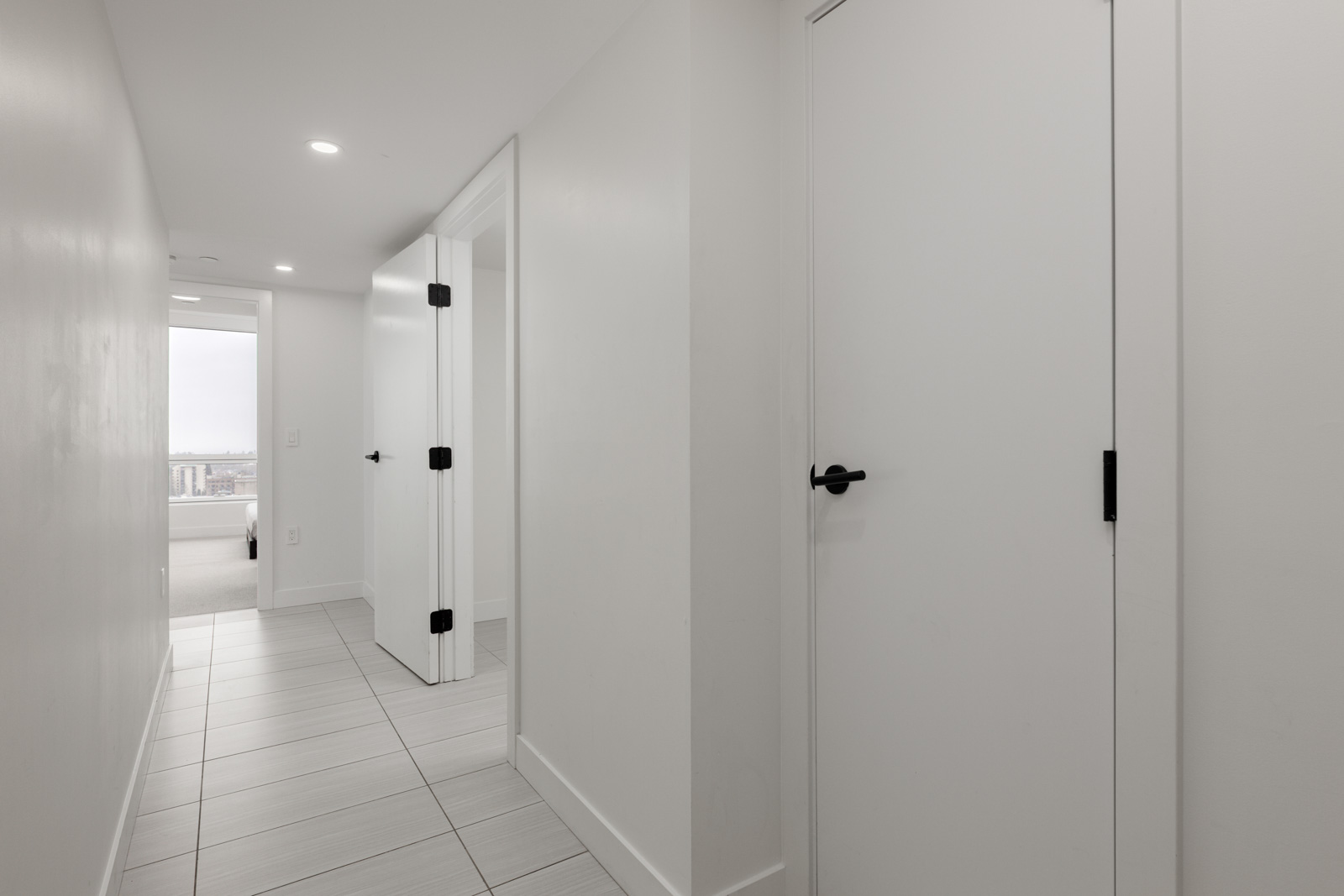 A modern, white hallway with tiled floors, closed doors on each side, and natural light coming from a window at the far end.