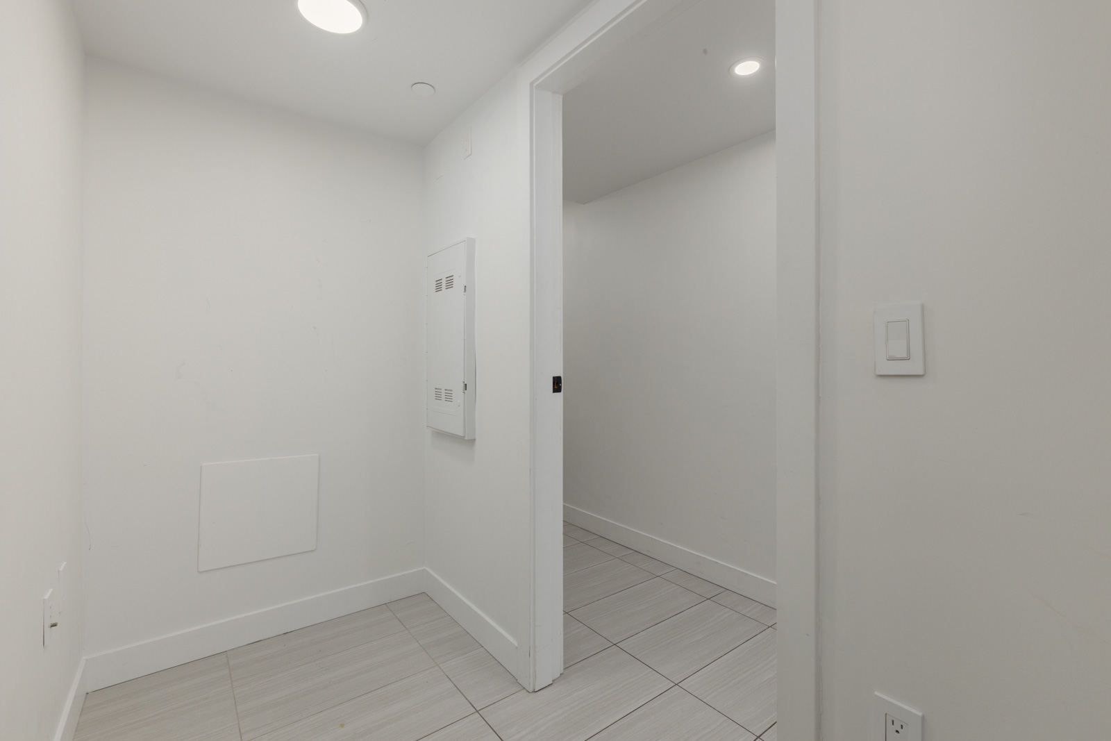 A small, empty white room with tiled floor, electrical panels on the wall, and an open doorway leading to another similar room.