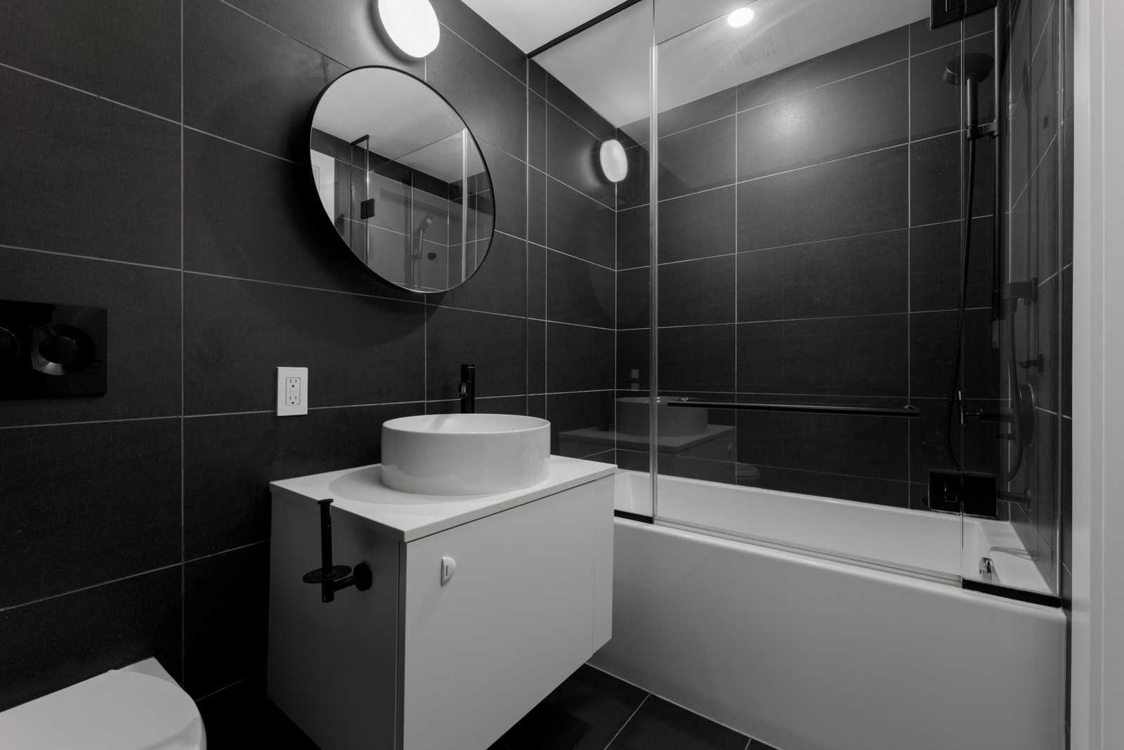 Modern bathroom with dark tiles, a round mirror above a white sink on a floating vanity, a bathtub with glass shower screen, and built-in lighting.