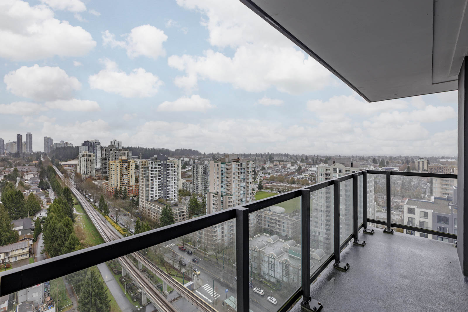 High-rise balcony with glass railing overlooking a cityscape of residential buildings, a tree-lined street, and a rail line under a cloudy sky.
