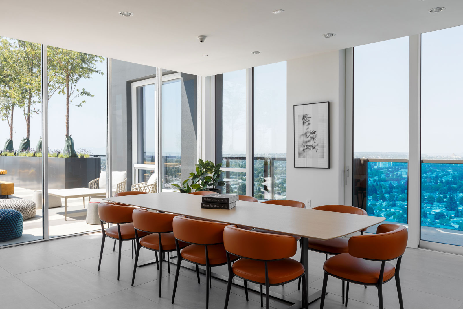 Modern dining room with a long table, eight brown chairs, potted plant, framed art on the wall, and large windows opening to a balcony with seating and city views.